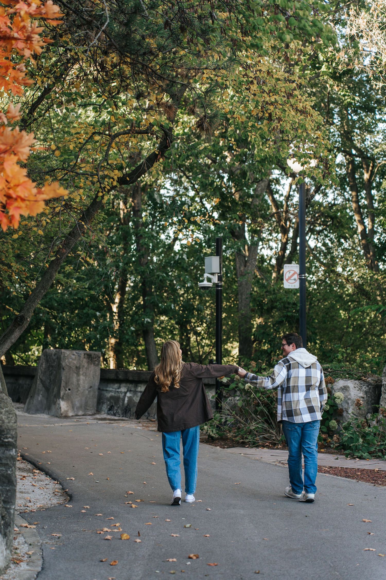 A person wearing jeans and plaid shirt walks down a tree-lined path in an autumn setting.