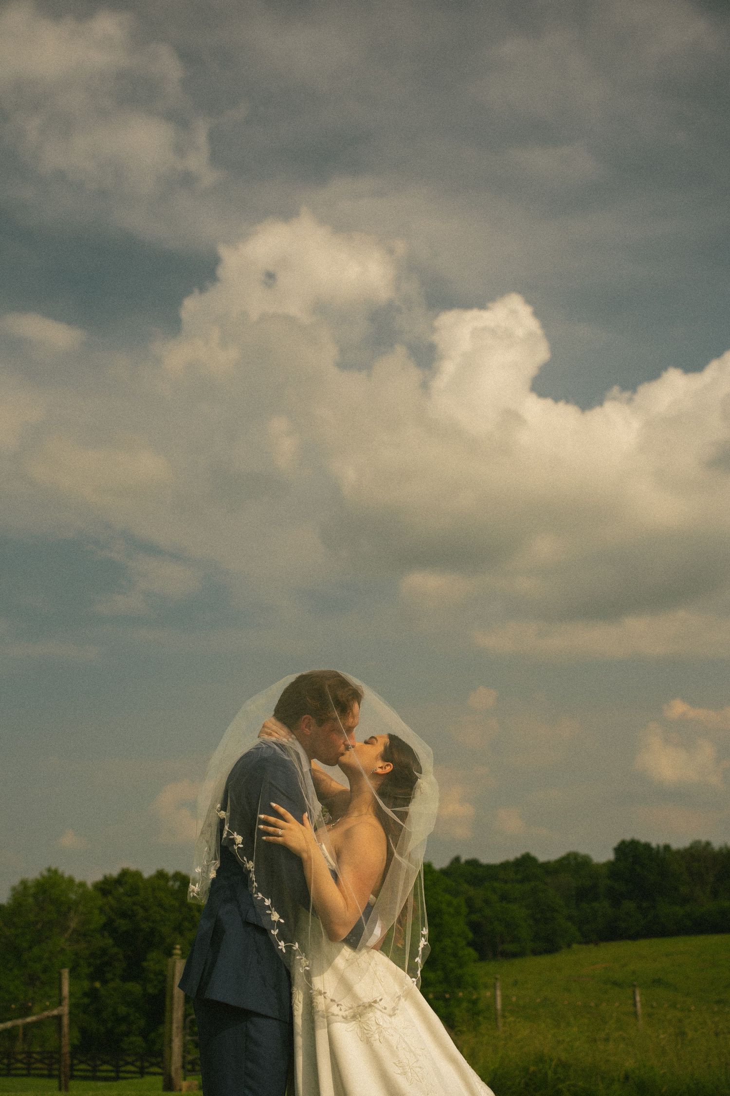 Bride and groom kissing under a flowing veil in a lush green field with dramatic cloudy sky backdrop.