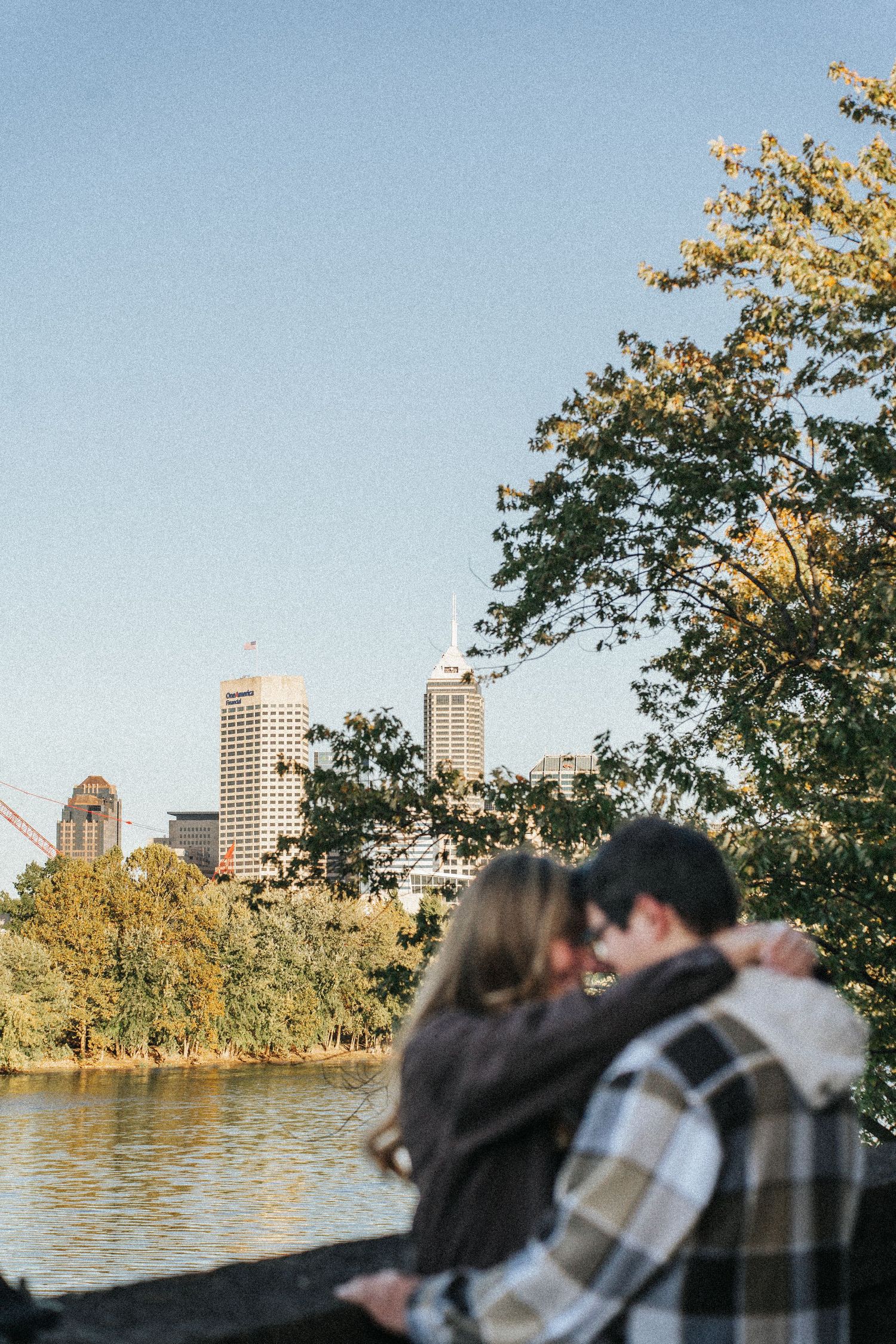 Couple embraces by a scenic lake with city buildings and autumn trees in the background during sunset.
