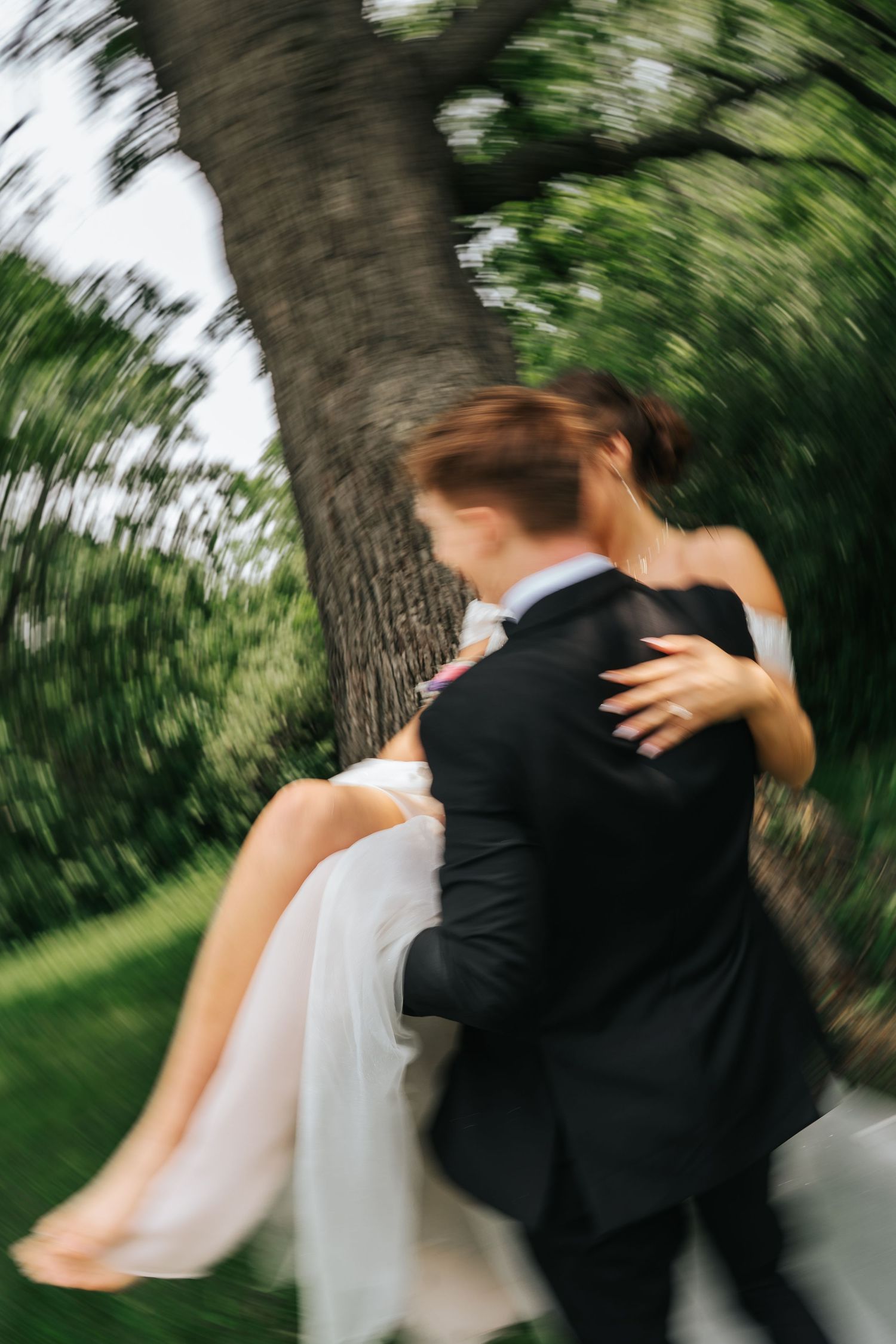 A romantic wedding moment captured in motion as a couple embraces near a tree in an outdoor garden setting.