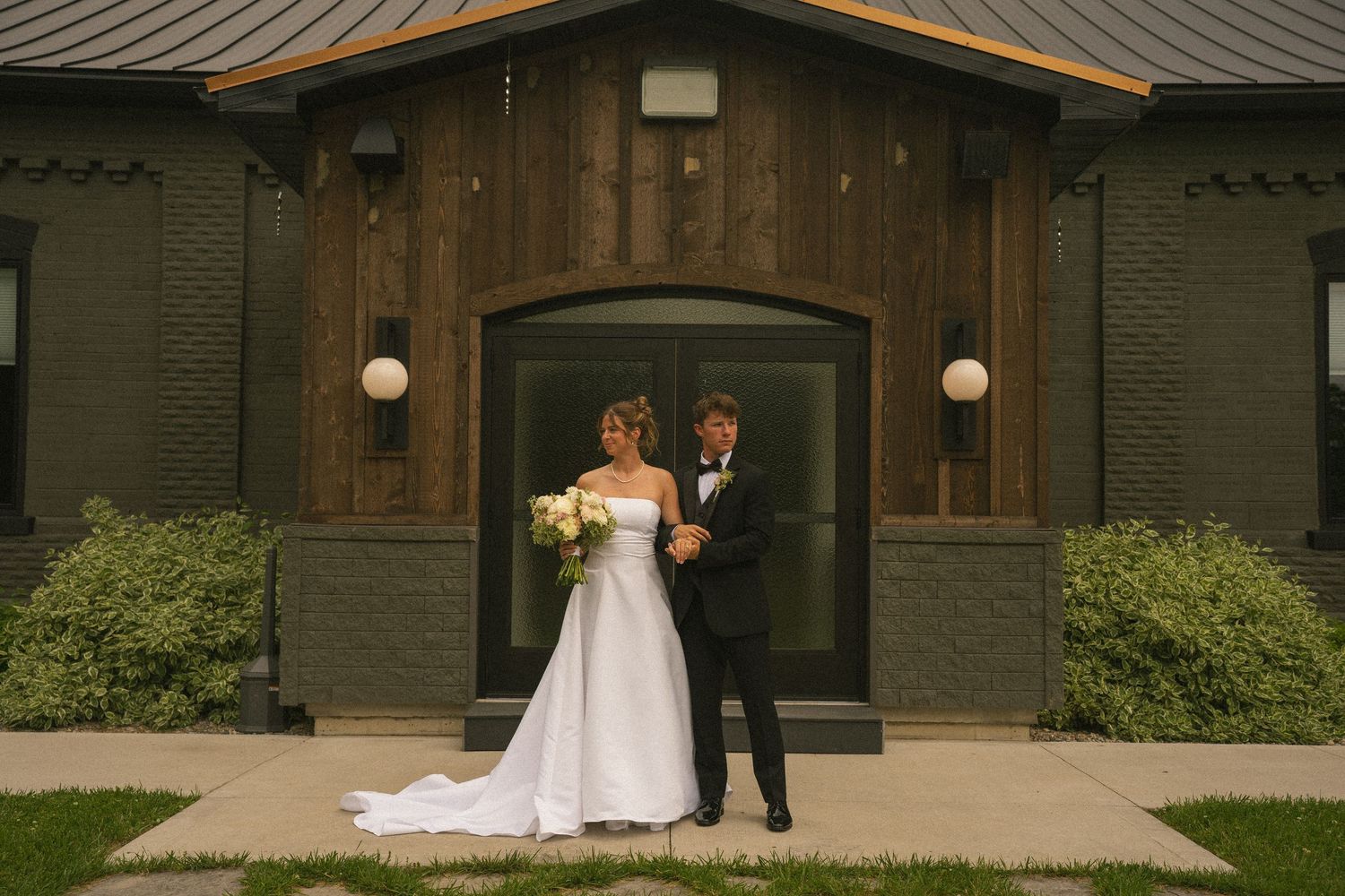 Bride and groom pose together outside a dark brick wedding venue with wood accents, bride holding white bouquet.
