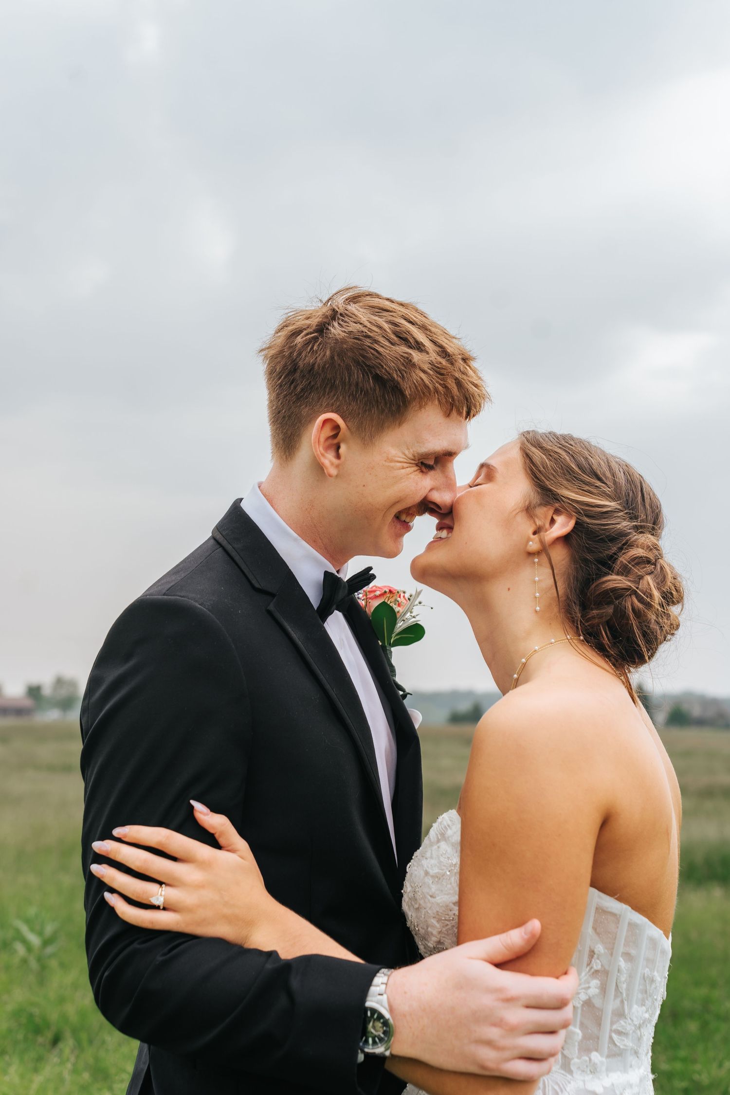 A romantic wedding couple embraces in an outdoor field setting with cloudy skies in the background.