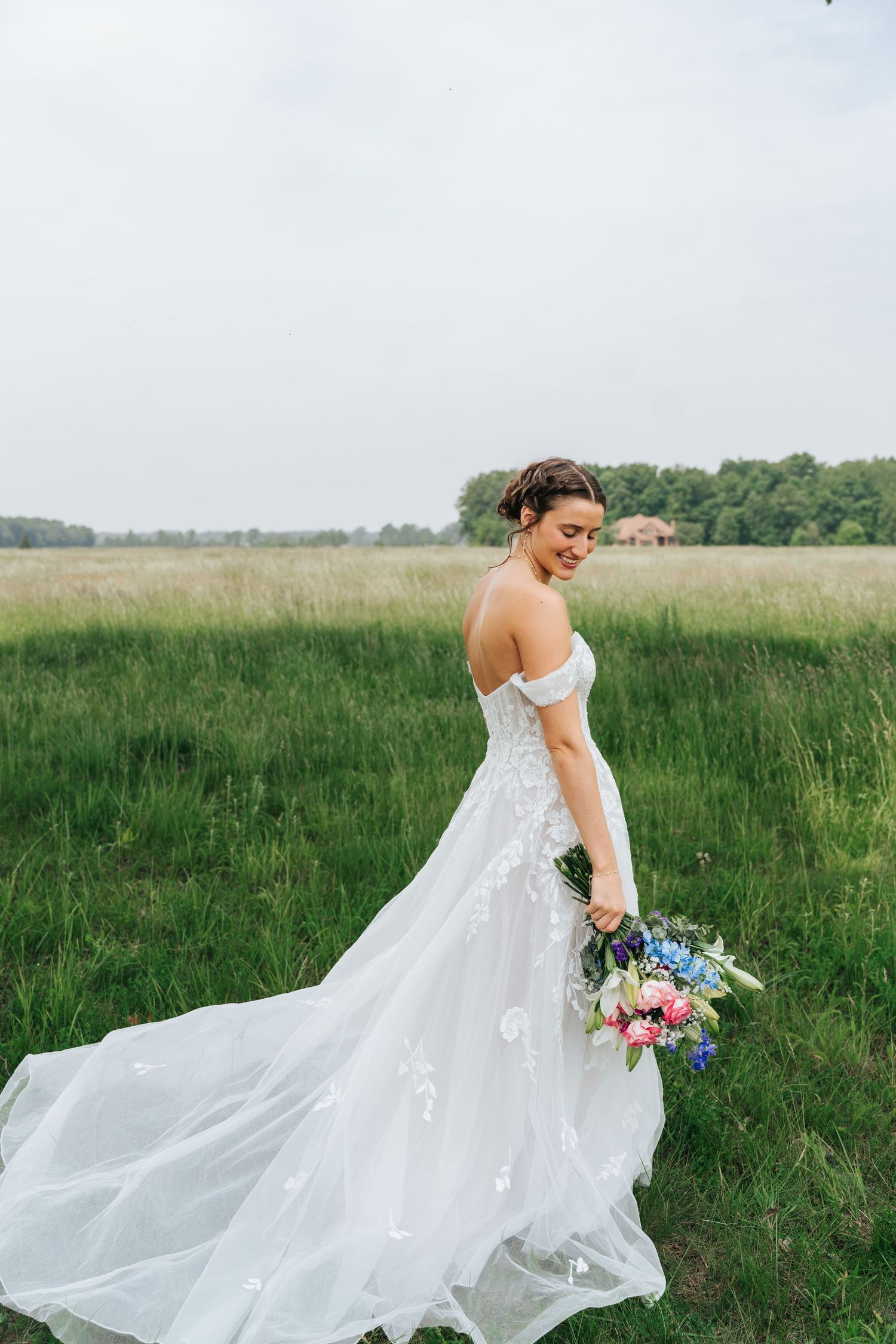 A bride in a flowing white lace wedding gown holds a colorful bouquet while standing in a serene meadow.