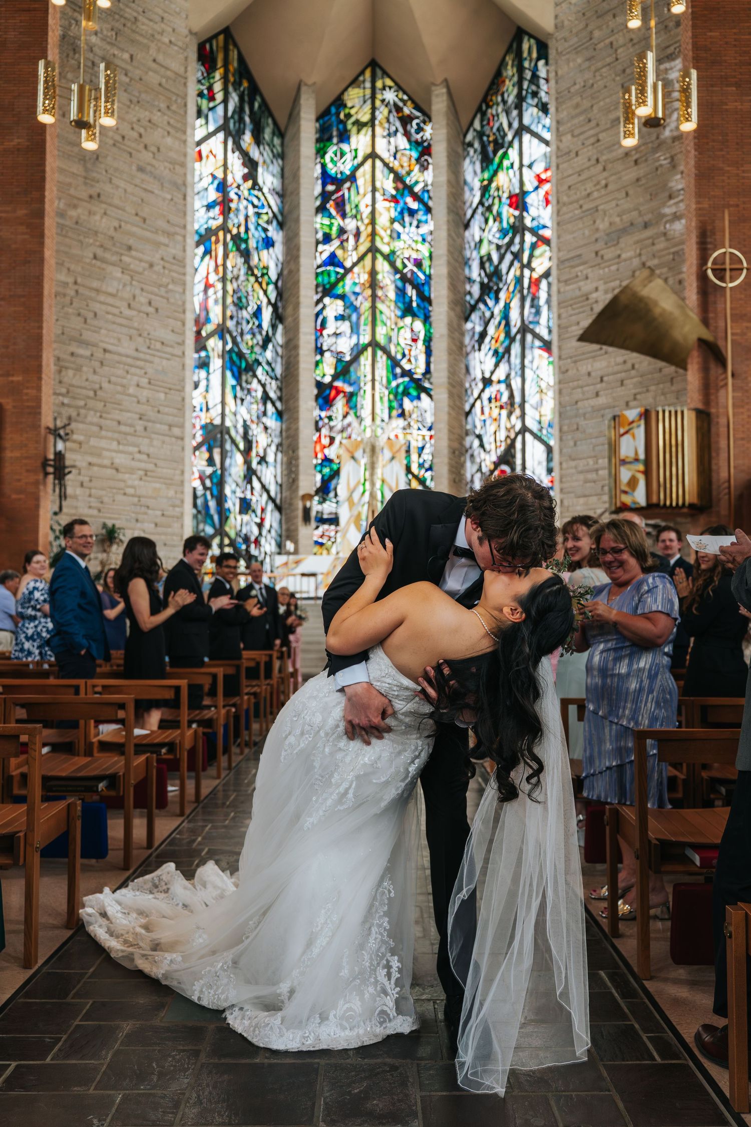 A couple shares their first kiss during a wedding ceremony in a church with stunning stained glass windows.