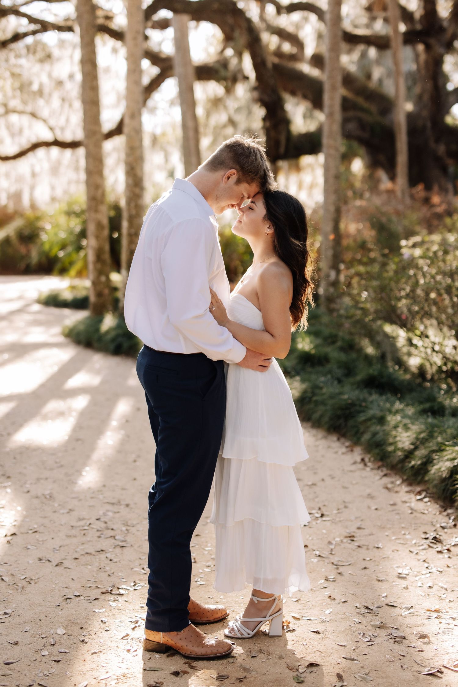 Couple sharing a romantic moment on a sunlit path, woman in white dress, man in white shirt and navy pants.