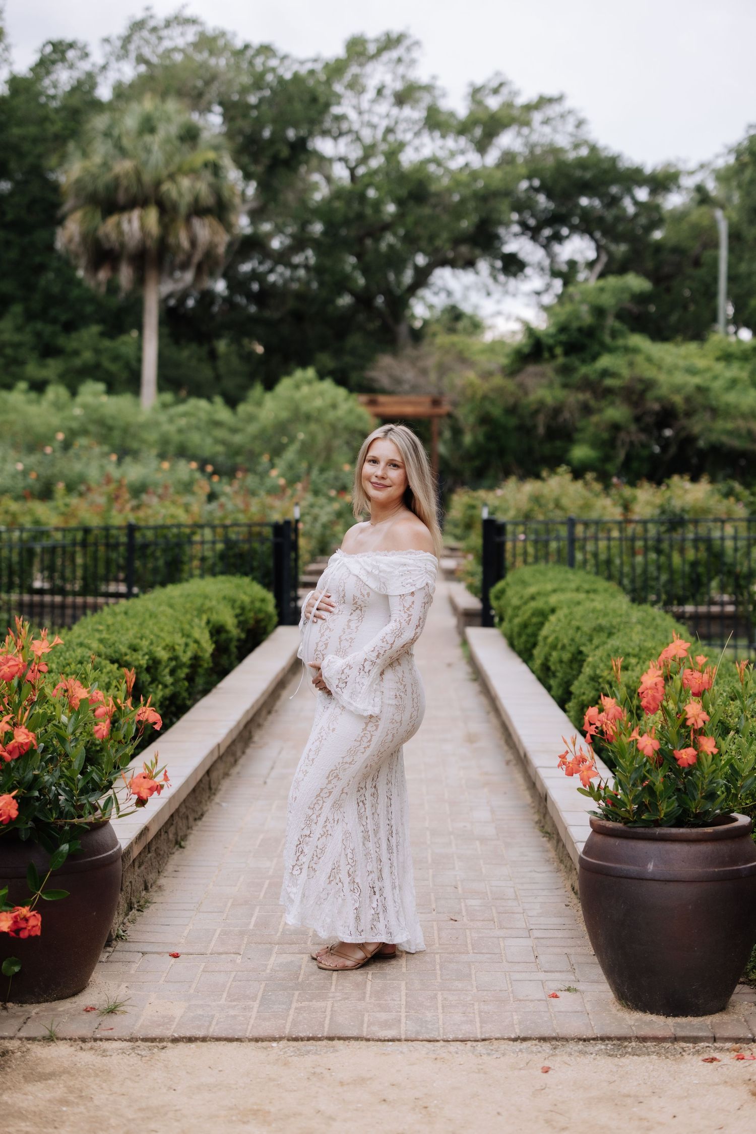 Pregnant woman in white lace off-shoulder gown poses in lush garden with orange flowers and manicured hedges.