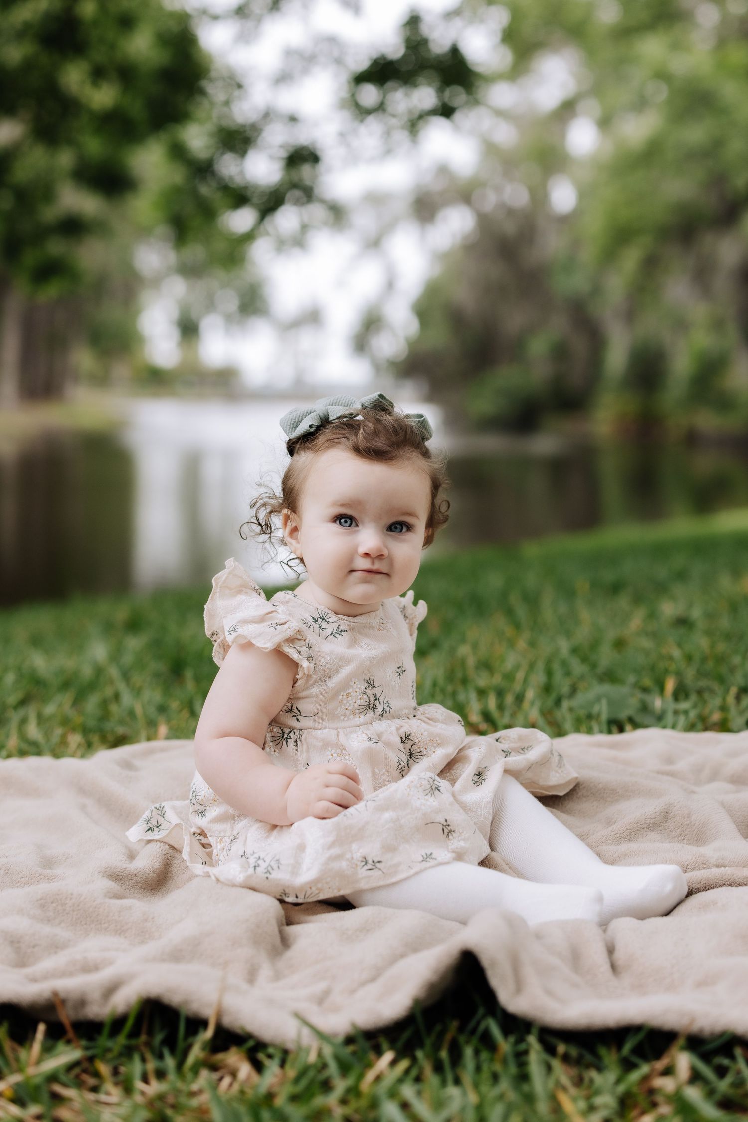 A baby girl in a floral dress sits on a blanket in a lush green park, looking curiously at the camera.