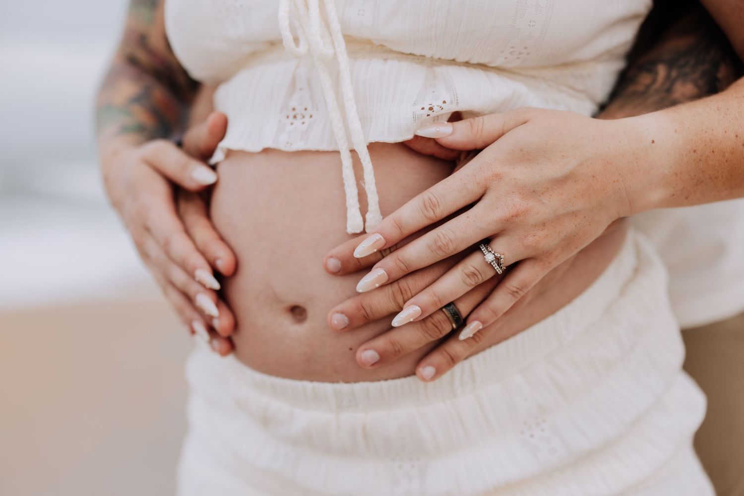 Pregnant woman in white crop top with tattooed arms, both partners hands cradling baby bump at beach.