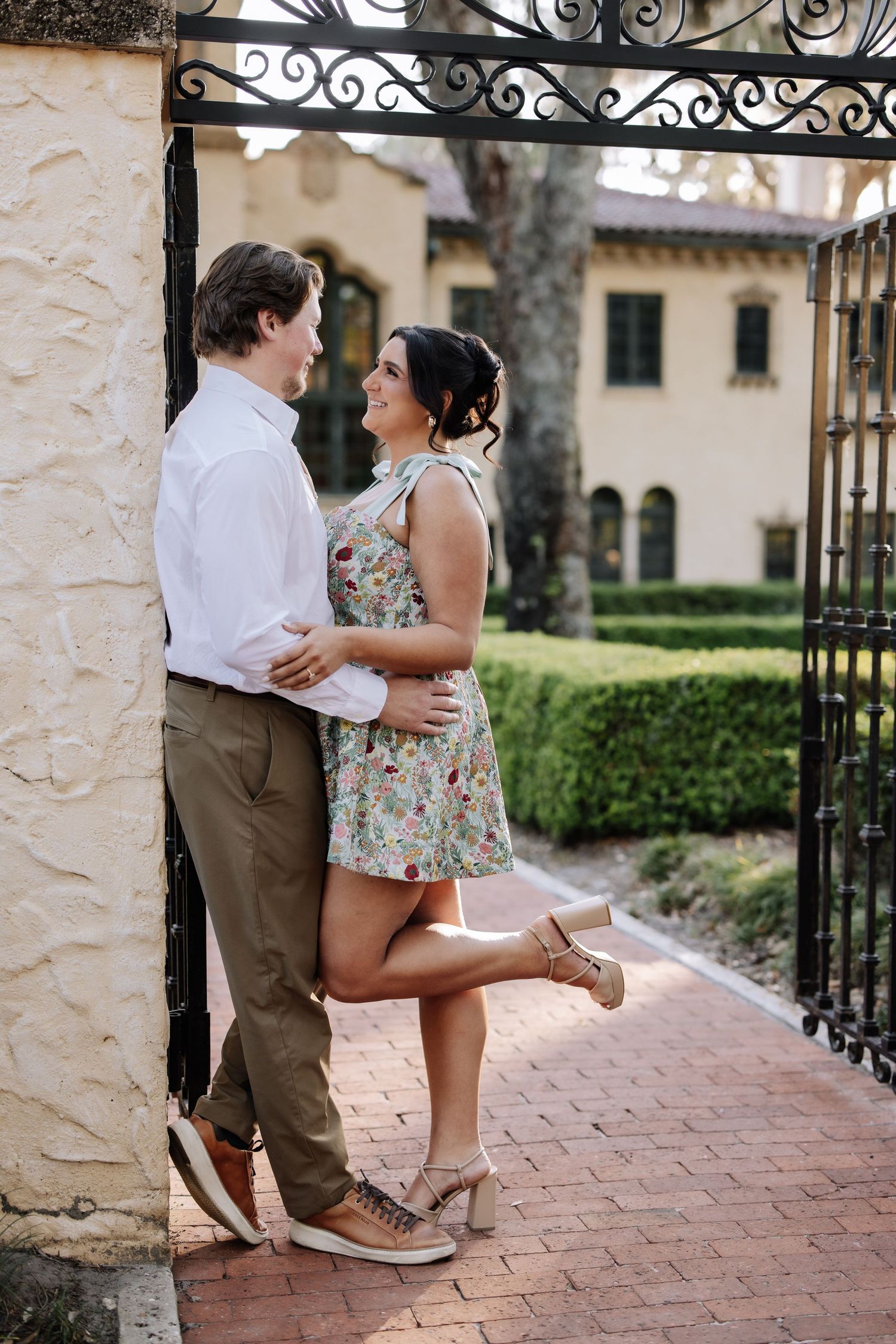 Couple embracing and smiling at each other by ornate iron gate, woman in floral dress, man in white shirt.