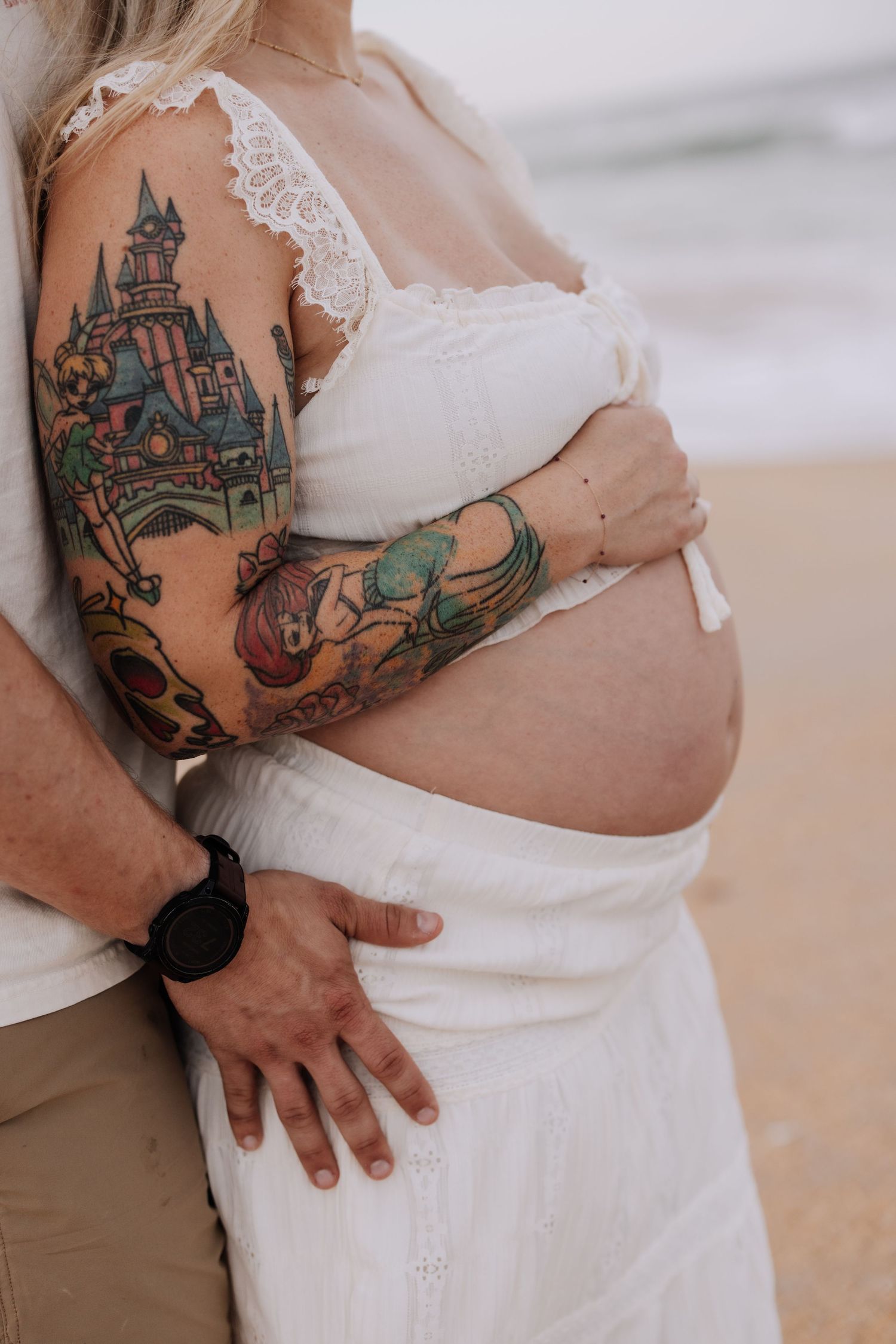 Pregnant woman in white dress with colorful tattoos embraced by partner on beach.