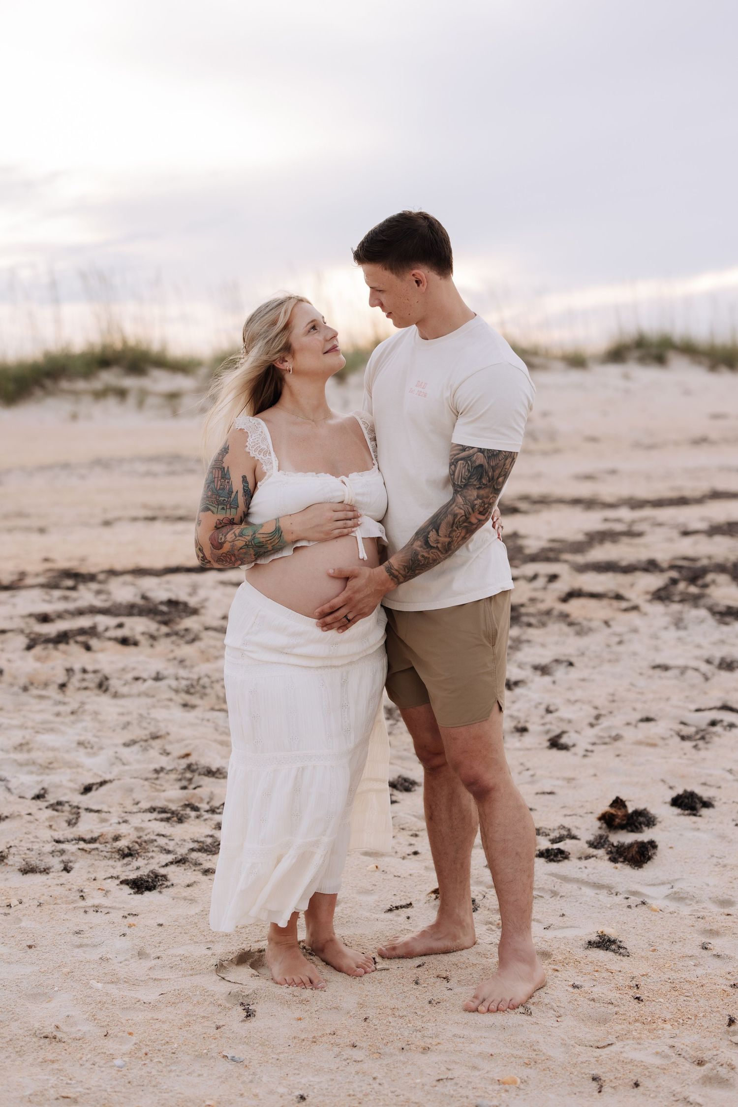 Tattooed couple sharing a tender moment on a sandy beach, woman in white dress cradling baby bump at sunset.