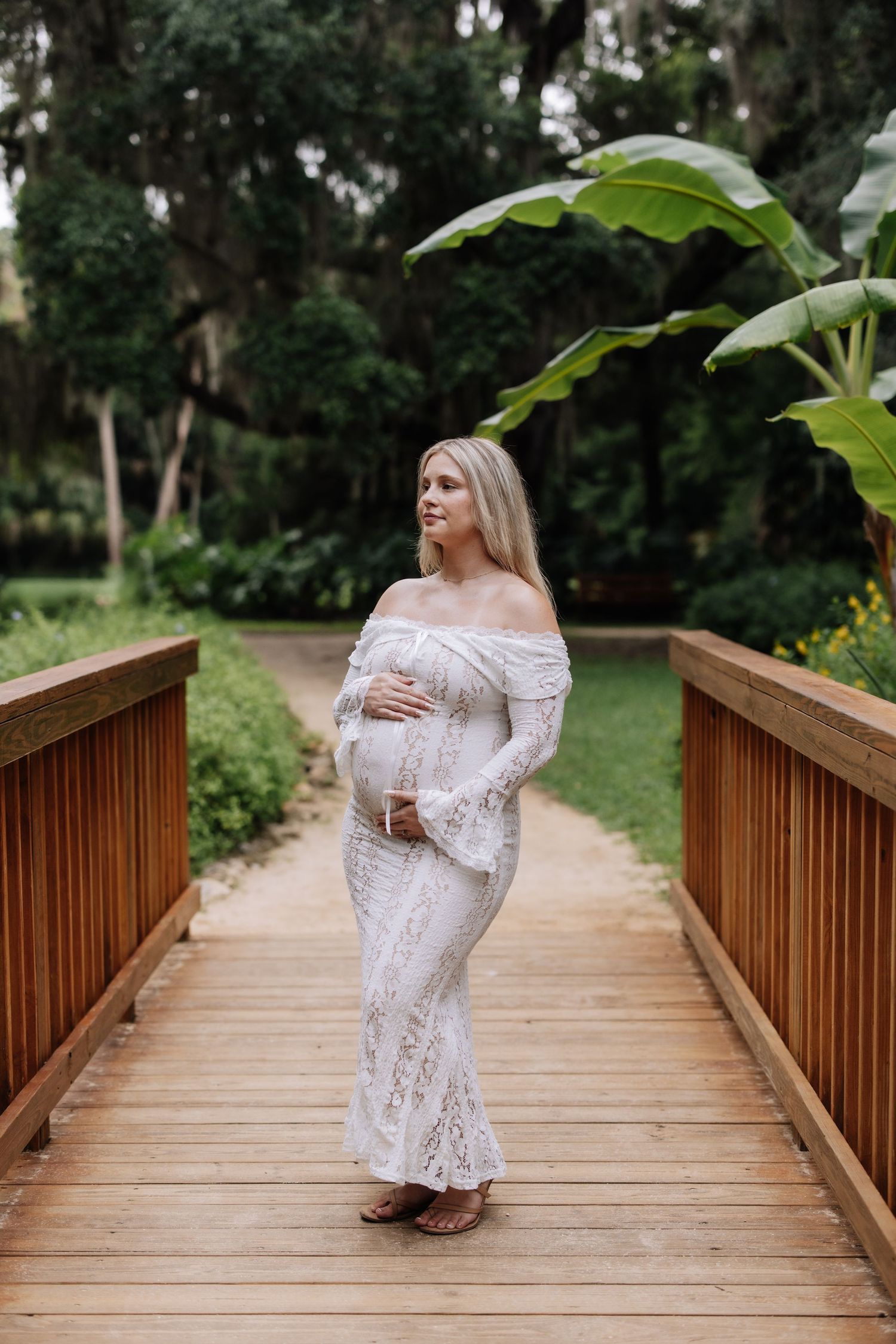 Pregnant woman in white lace off-shoulder gown on wooden bridge surrounded by lush tropical greenery.