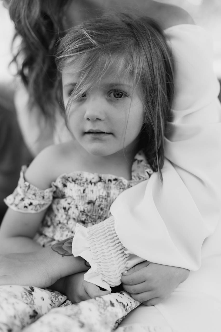 Black and white portrait of a young girl in a floral off-shoulder dress, held by an adult in white clothing.