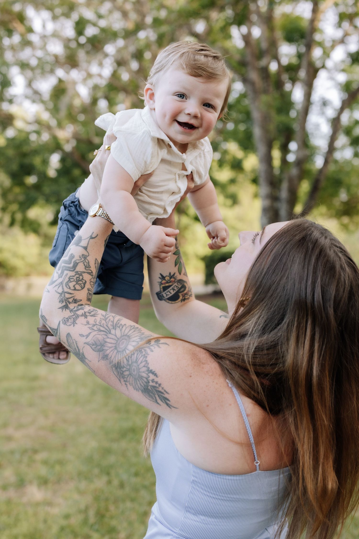 Tattooed mother joyfully lifting laughing baby in the air outdoors in a lush green park setting.