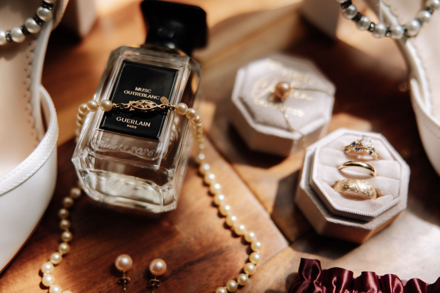Givenchy perfume bottle with pearl necklace, gold rings in velvet boxes, and white heels on wooden surface.