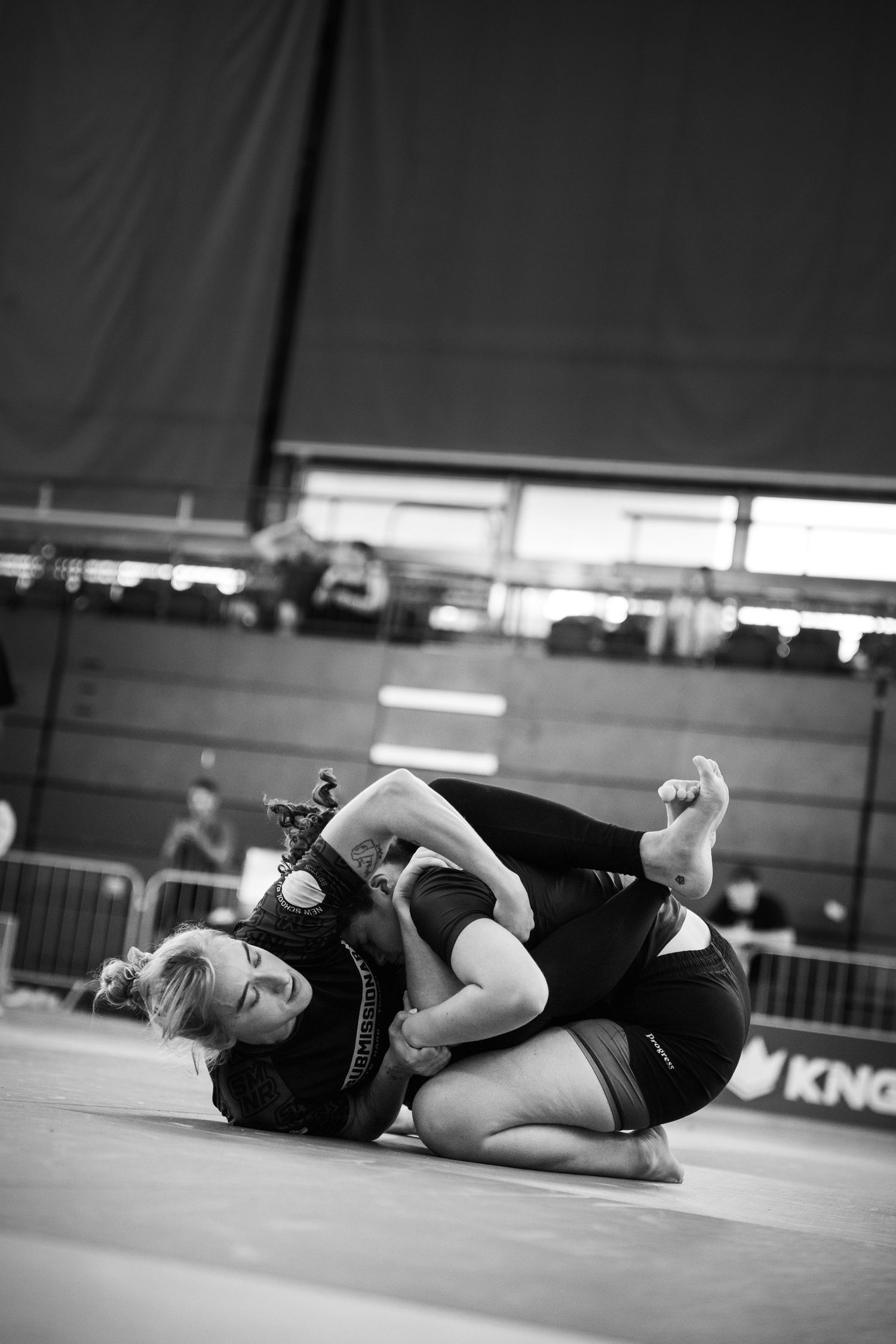 Two women competing in a Brazilian jiu-jitsu grappling match on a mat at an indoor tournament.