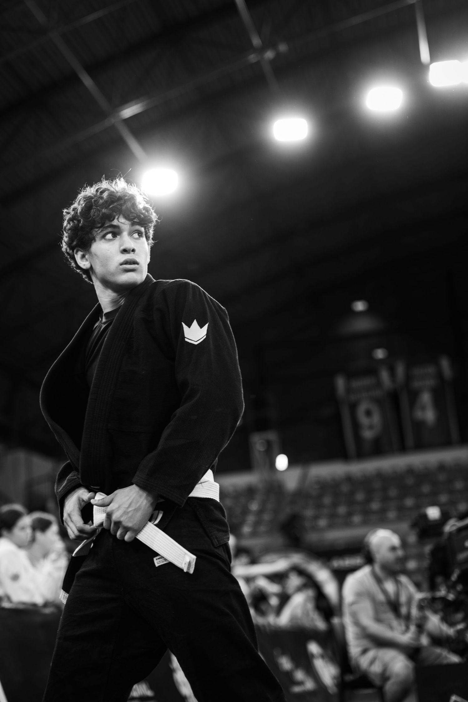 Young male BJJ competitor in black gi stands alert at a martial arts tournament in black and white photo.
