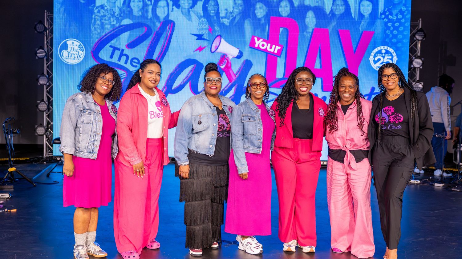 People wearing bright pink and blue outfits stand together at an event with colorful backdrop.