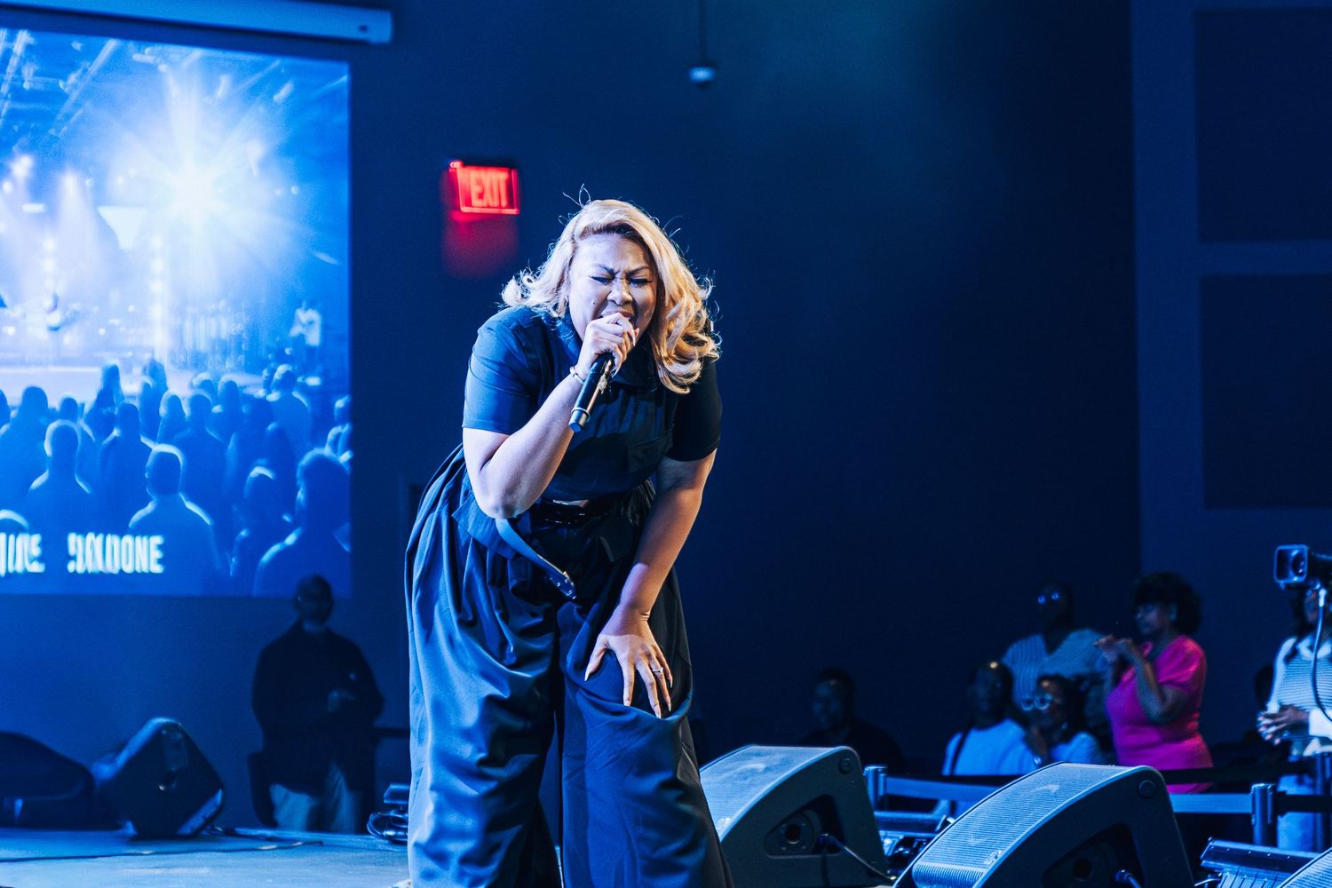 A performer sings passionately on stage with blue lighting and silhouetted crowd in the background.