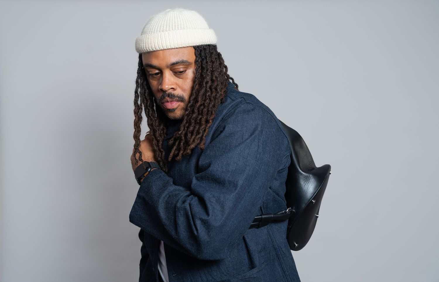 A man with dreadlocks wearing a white beanie and navy jacket holds a black bag against a gray background.