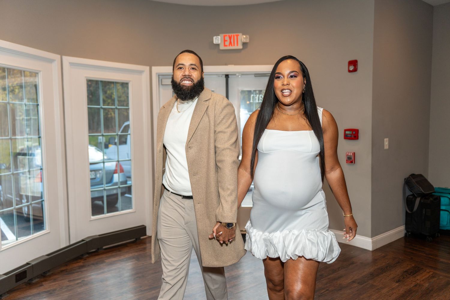 A couple holds hands while walking down a hallway, with the person on the right wearing a white dress.