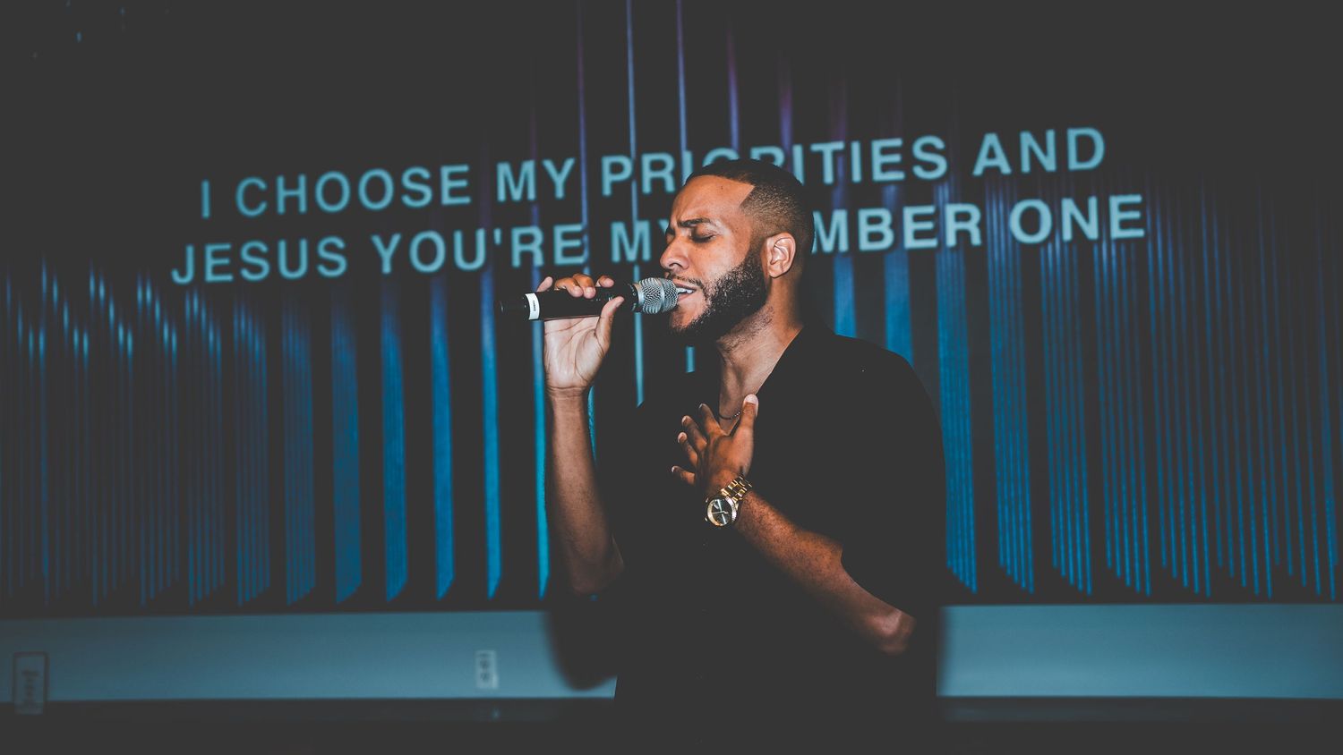 Stage performance with lyrics projected on screen reading 'I choose my priorities' in blue lighting.