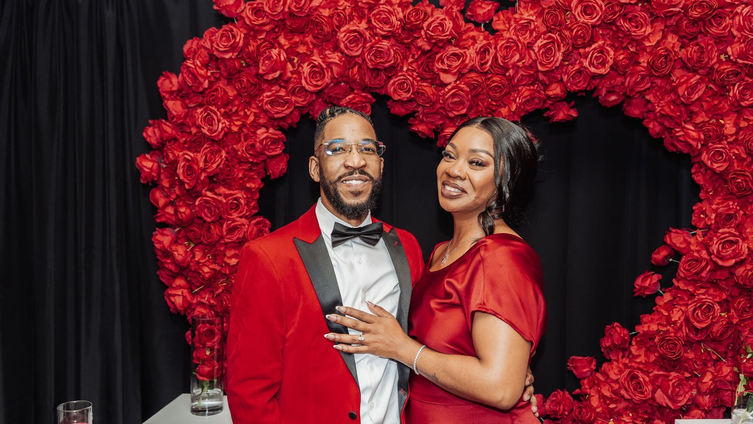 A couple in matching red attire pose together under a heart-shaped arch made of vibrant red roses.