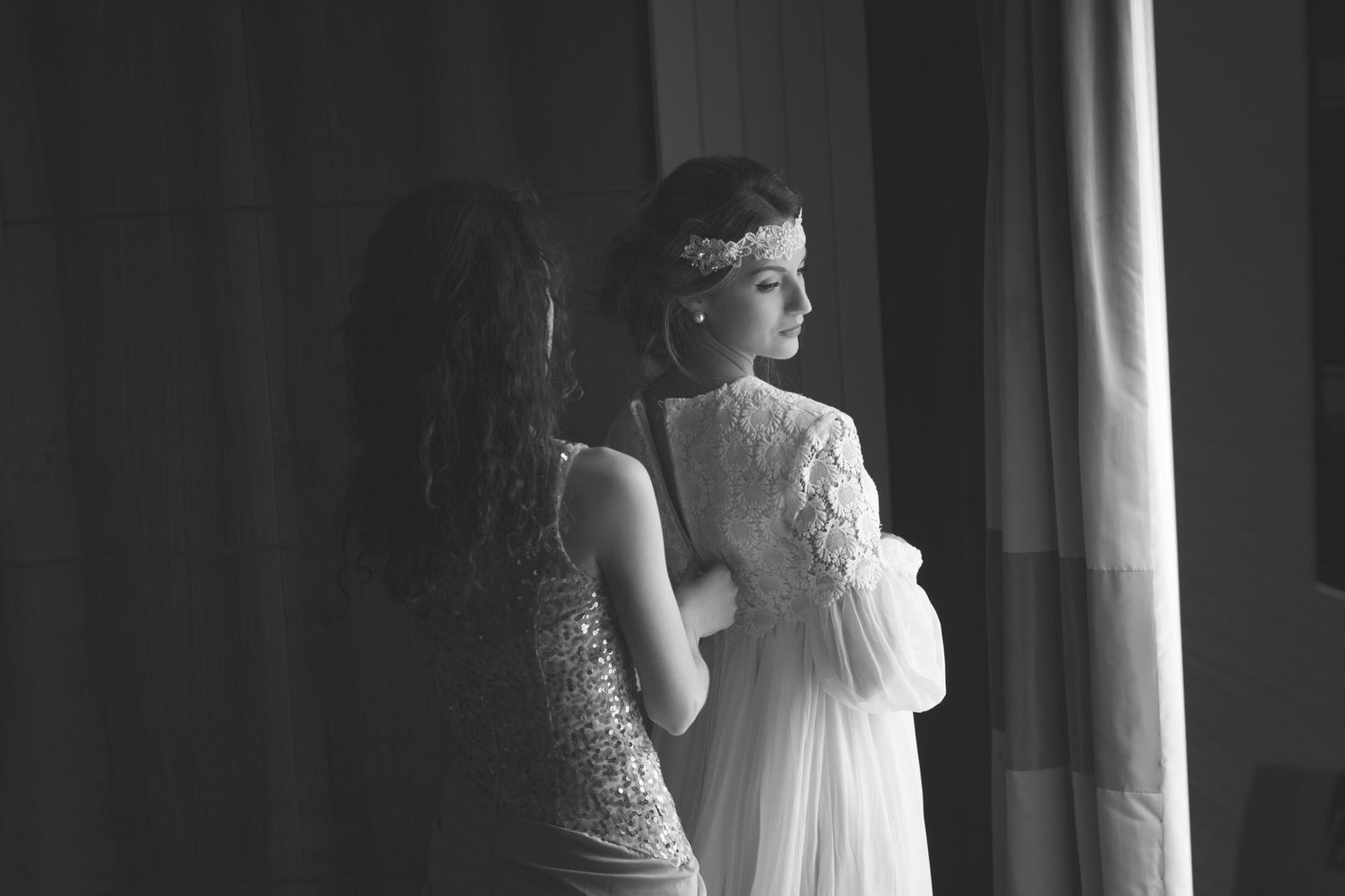 Bridesmaid helping bride with dress by window before wedding in Cork