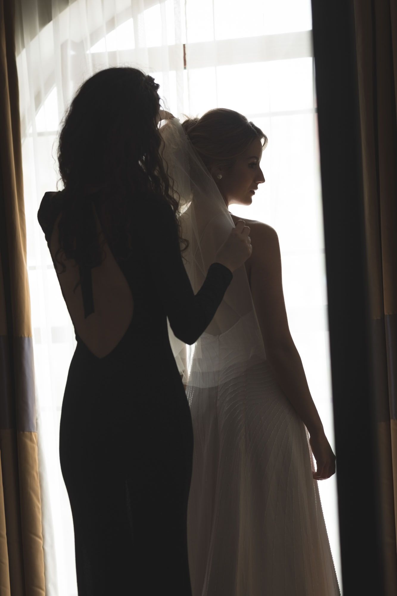 Bridesmaid helping bride with her veil by window before wedding in Cork
