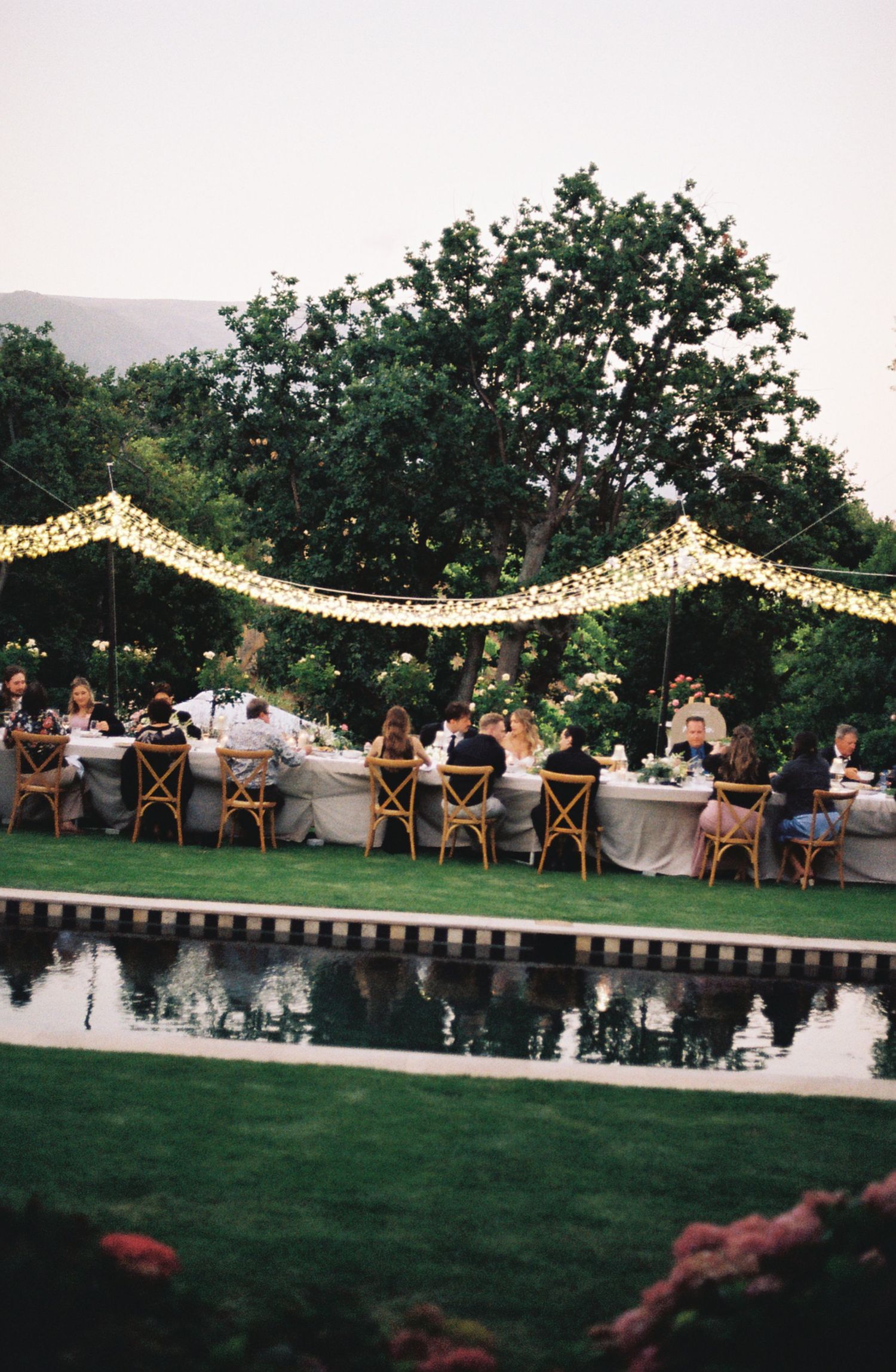 Wedding reception by a pool with string lights overhead and guests dining at long tables.