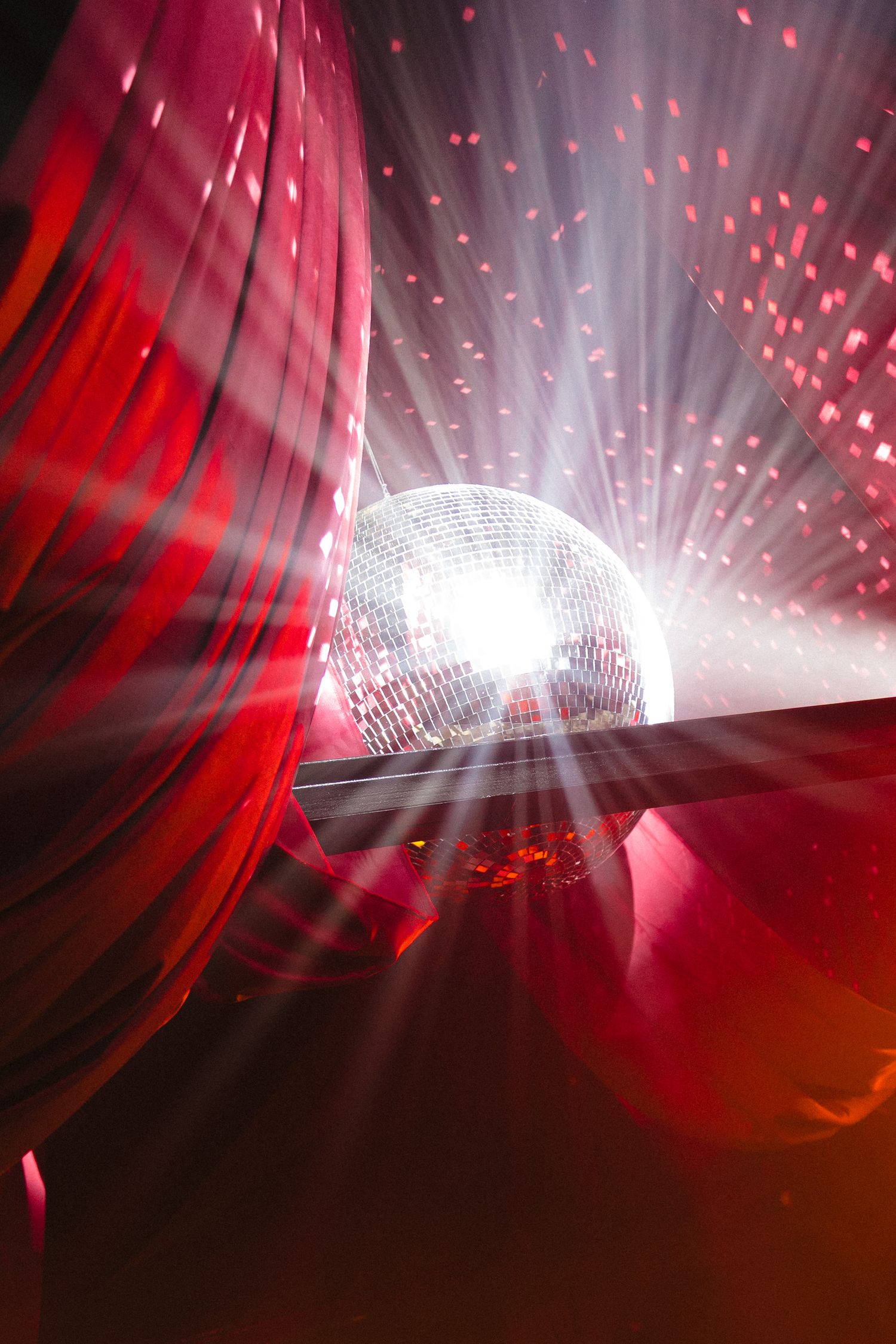 A glowing disco mirror ball casting red and white light rays through dramatic red curtains at a nightclub event.