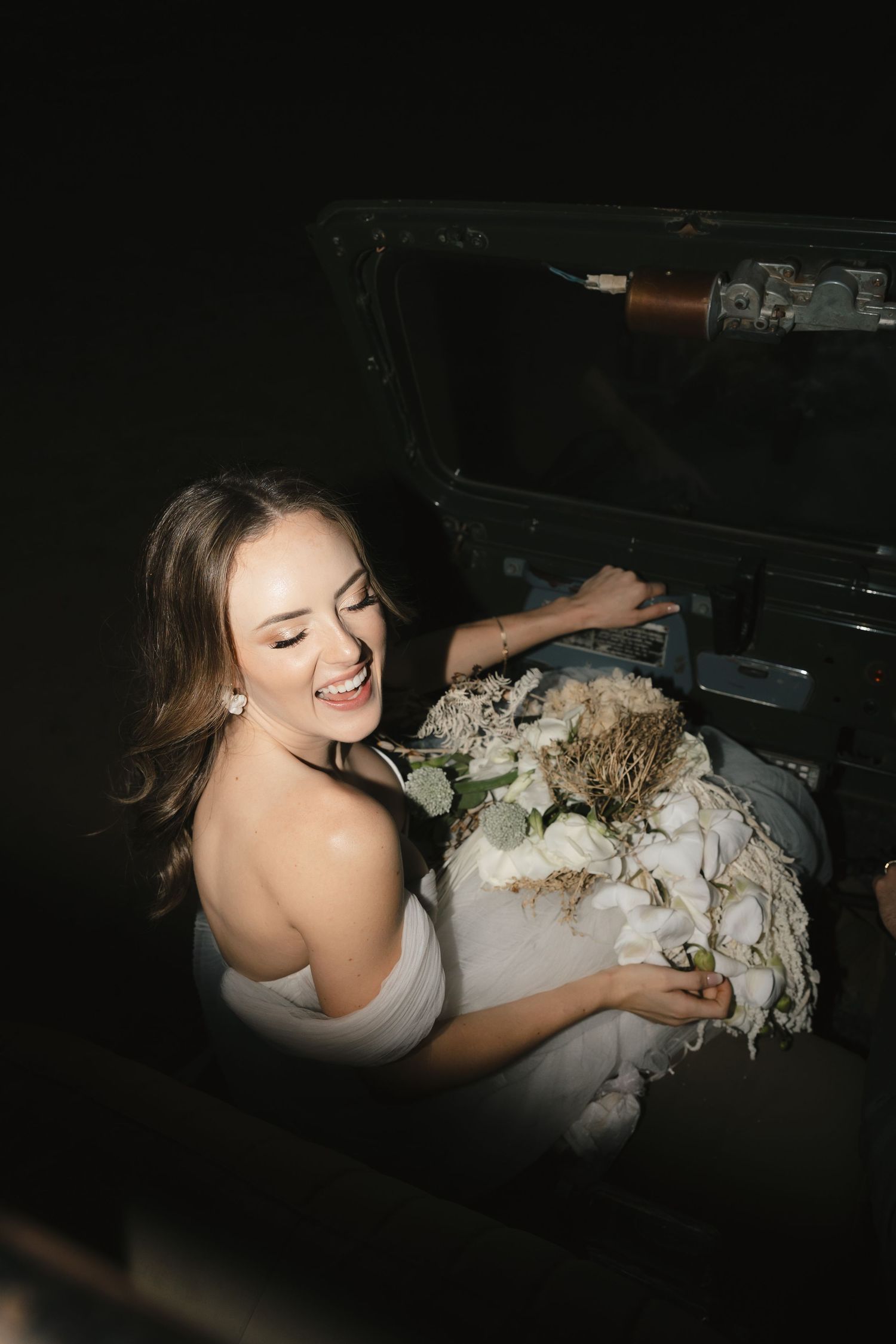 A person in a white dress lying on a dark surface while holding and touching what appears to be a floral arrangement.