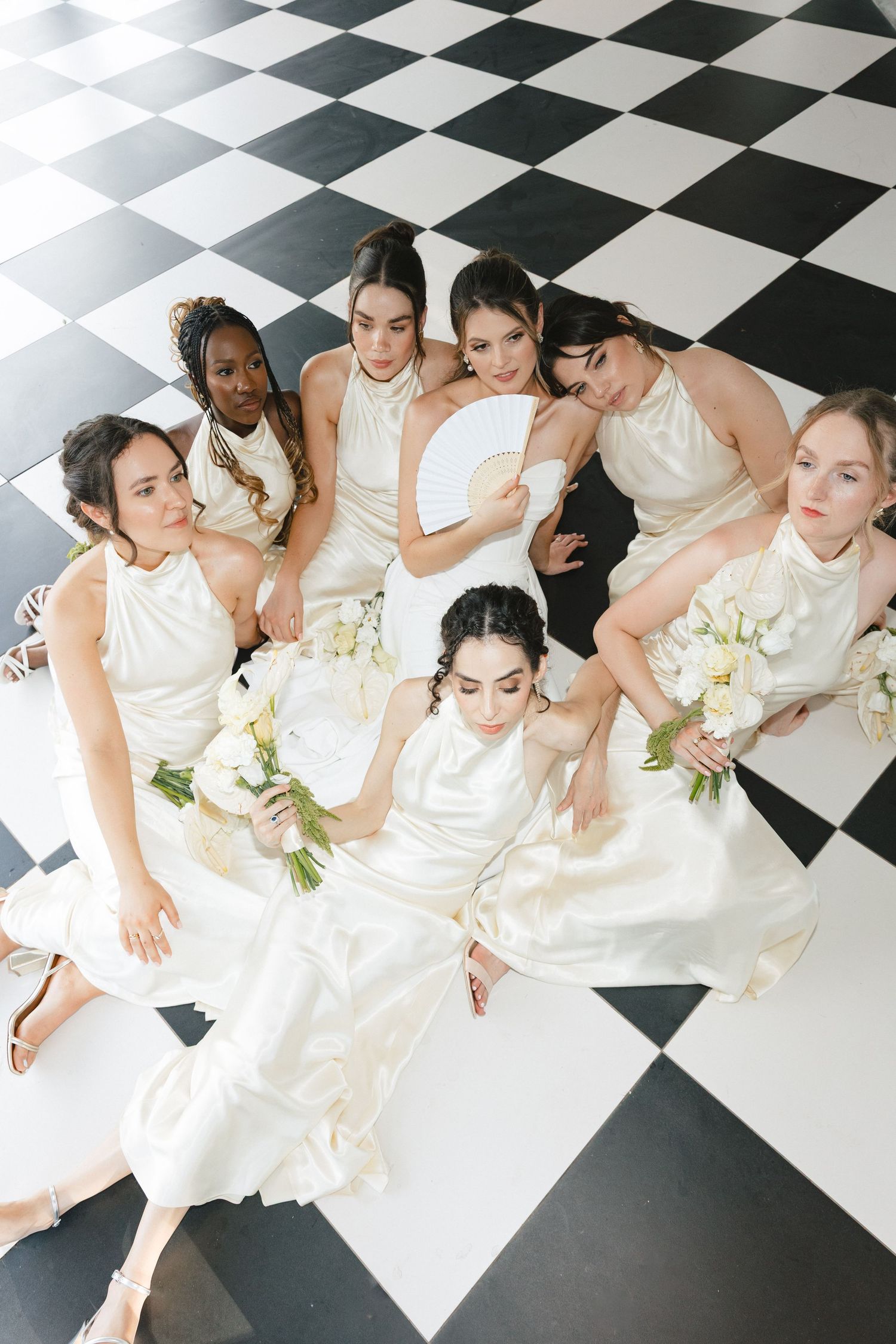 A group in white dresses pose together on a black and white checkered floor holding white bouquets.