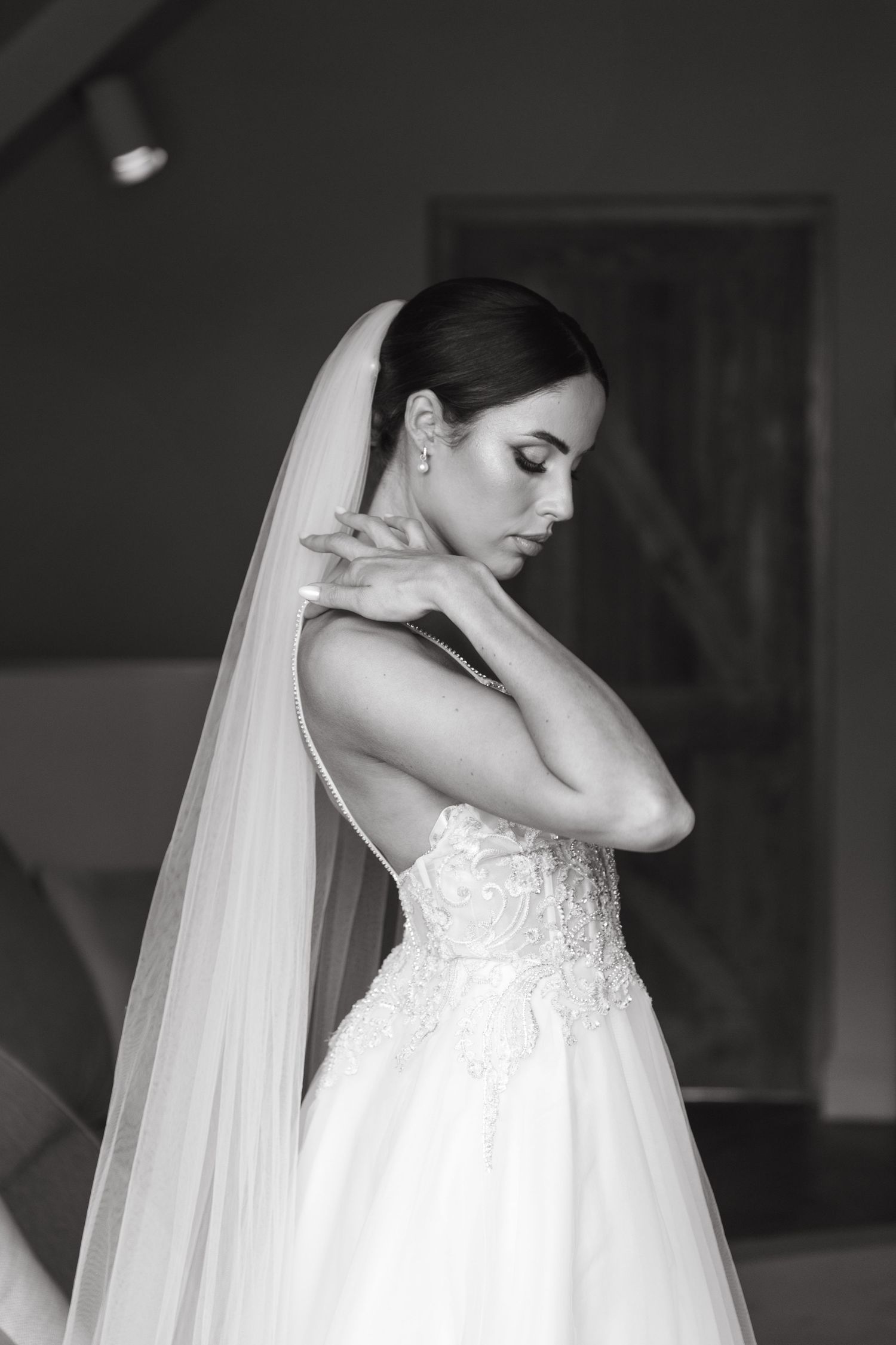 Black and white portrait of an elegant bride in a lace wedding gown and veil, posing gracefully indoors.