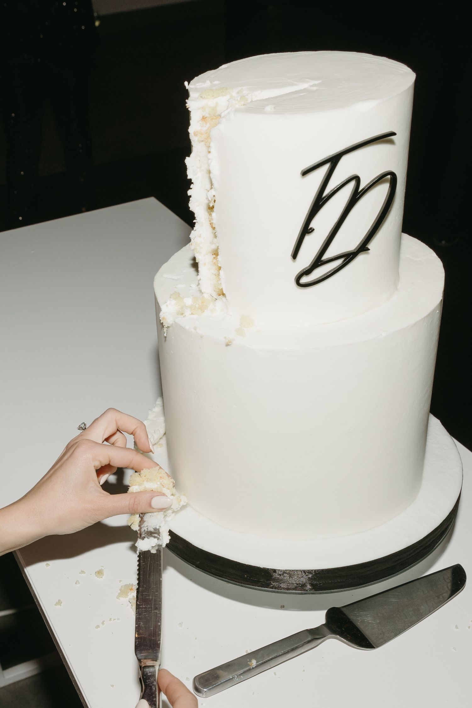 Two-tier white wedding cake with black monogram being cut, showing interior layers on a white table.