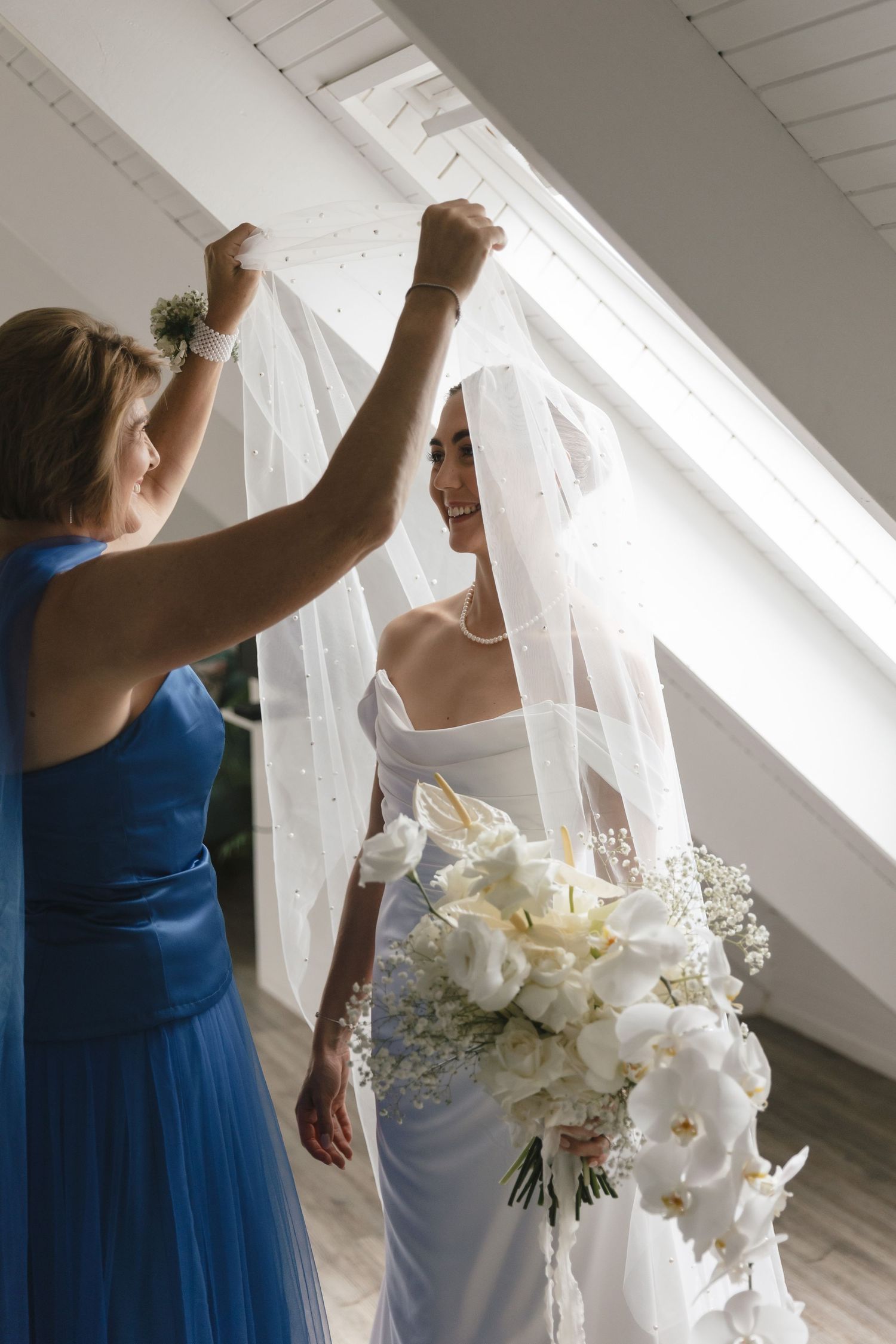A bridesmaid in blue helps adjust a flowing white wedding veil near a bright window.