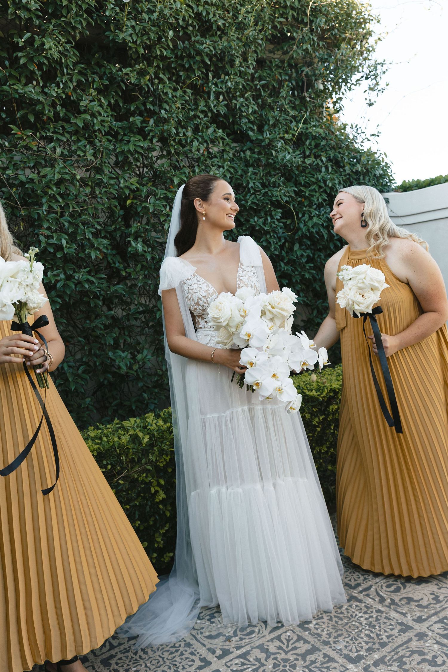 Two wedding guests in contrasting white and gold dresses share joyful candid moments against a green hedge backdrop.