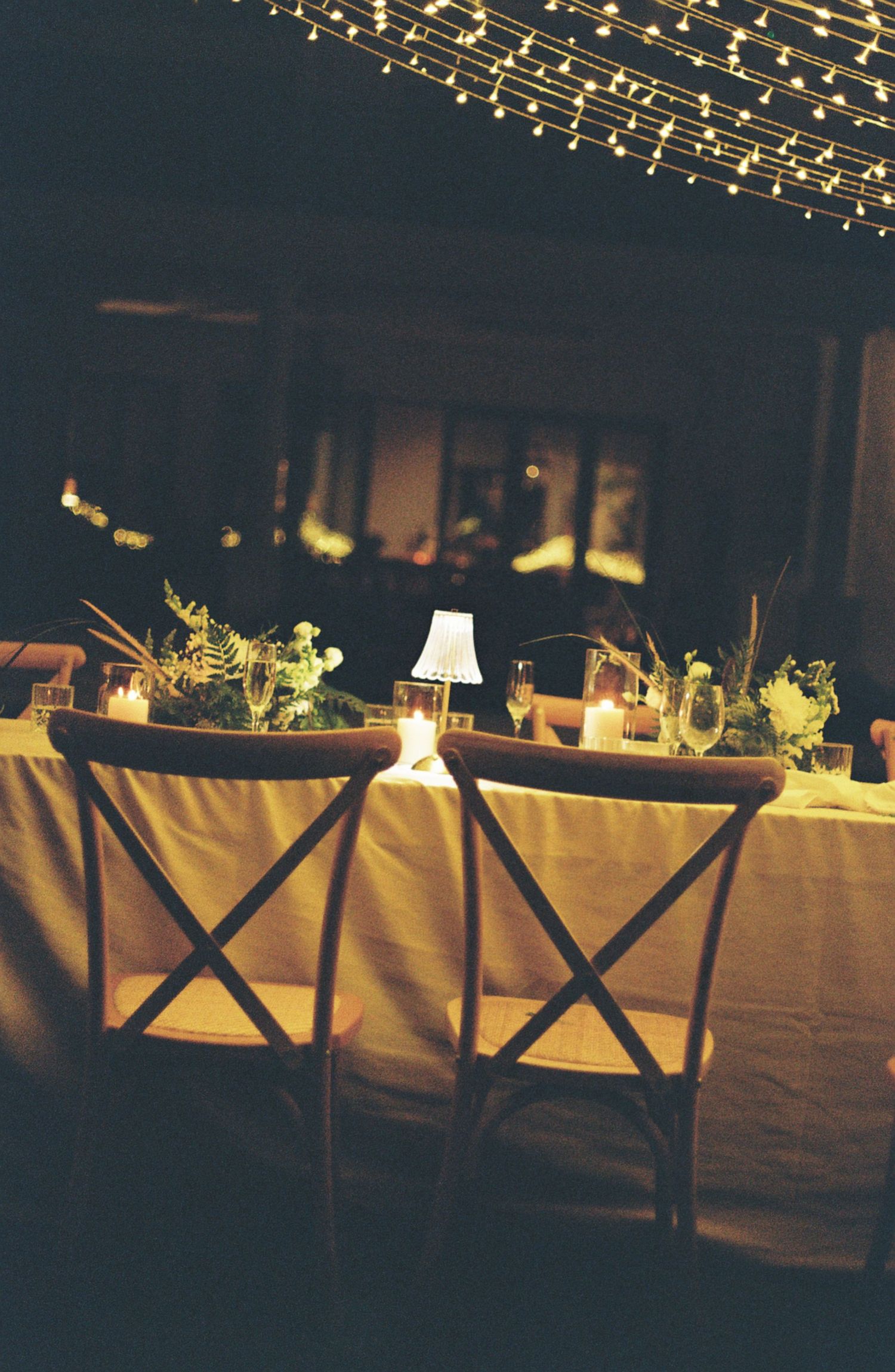 Rustic wooden chairs line a white-clothed table under twinkling string lights at an elegant evening reception.