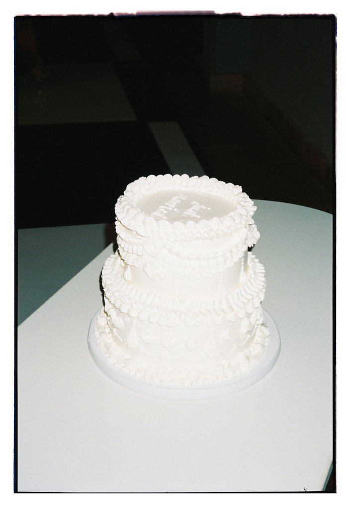 A white frosted wedding cake with delicate piping details sits elegantly on a table against a dark background.