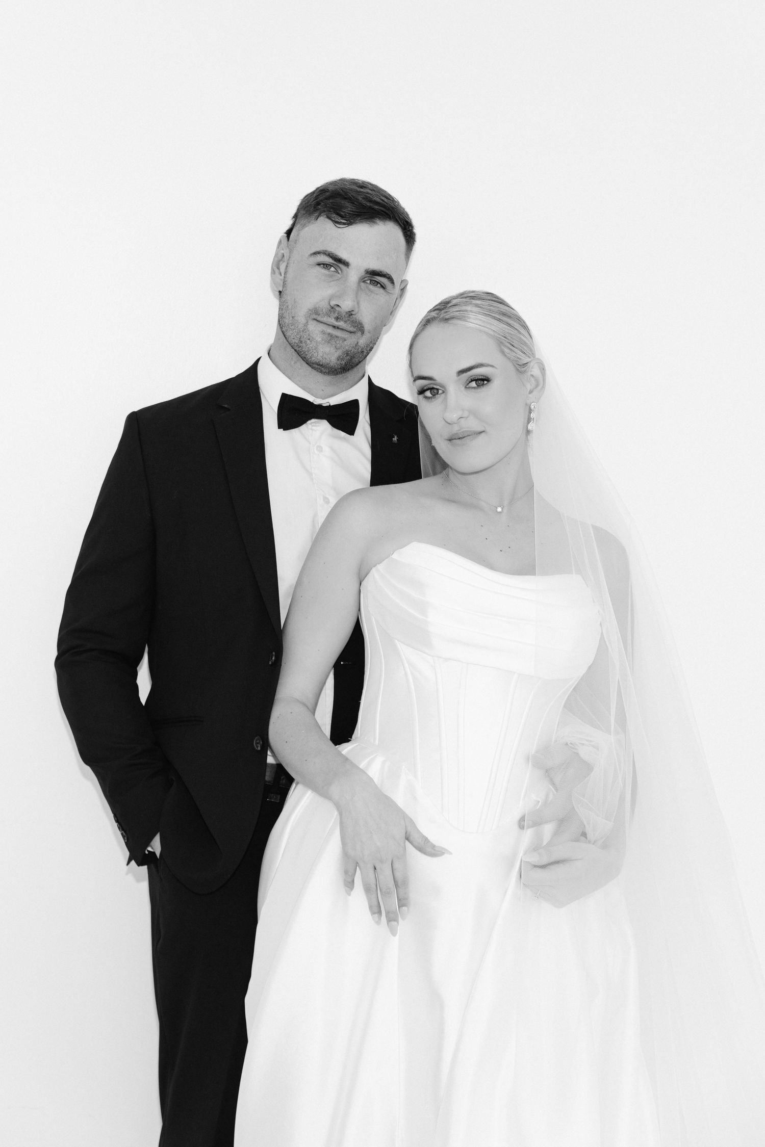 A couple poses together in formal black and white attire against a minimalist white studio backdrop.