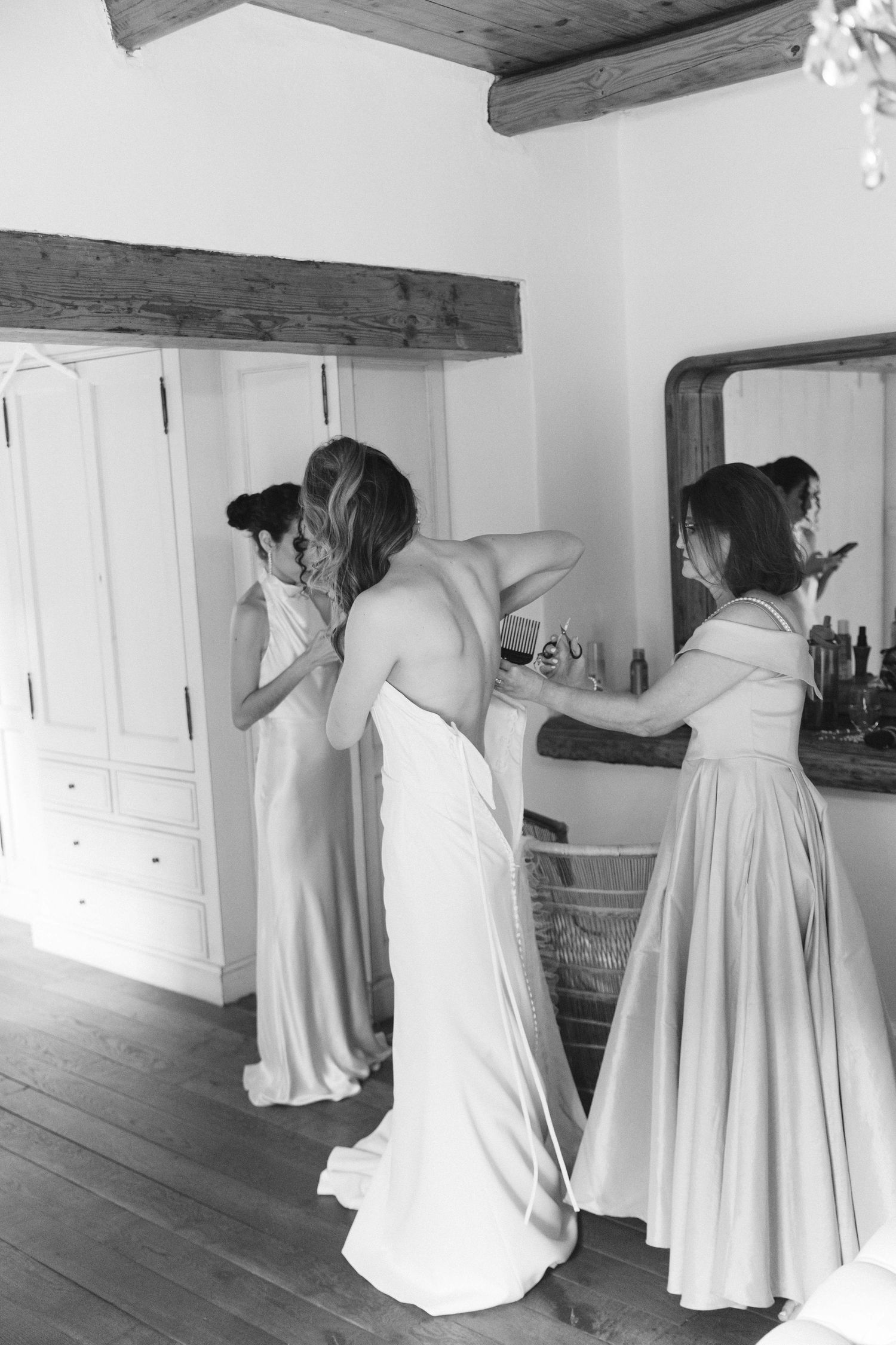 Wedding party getting ready in white dresses in rustic room with wooden beams.