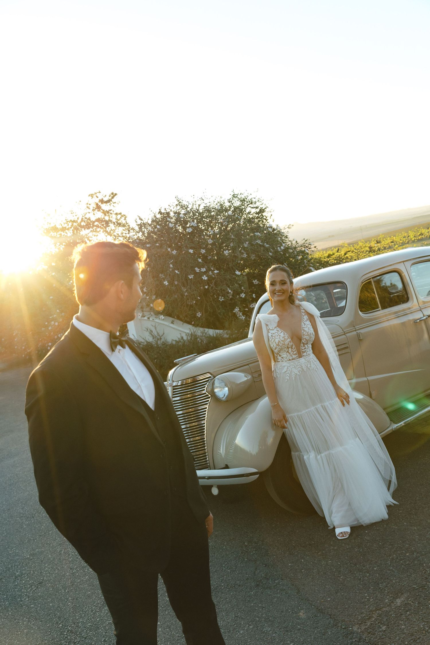 Vintage car parked at sunset serves as backdrop for elegant wedding photos with couple in formal attire.