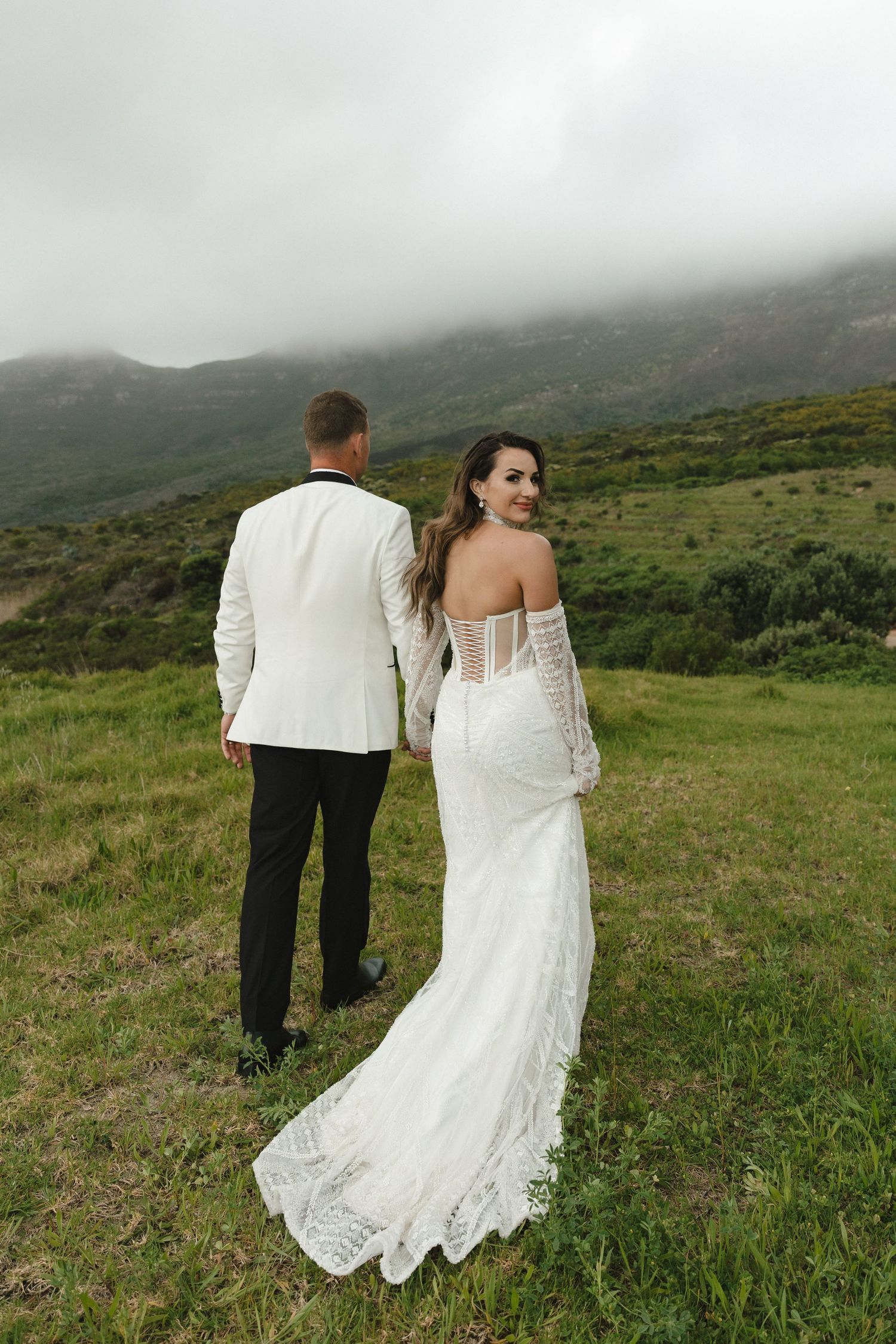A couple in wedding attire walks through a misty mountainside meadow with green rolling hills in the background.