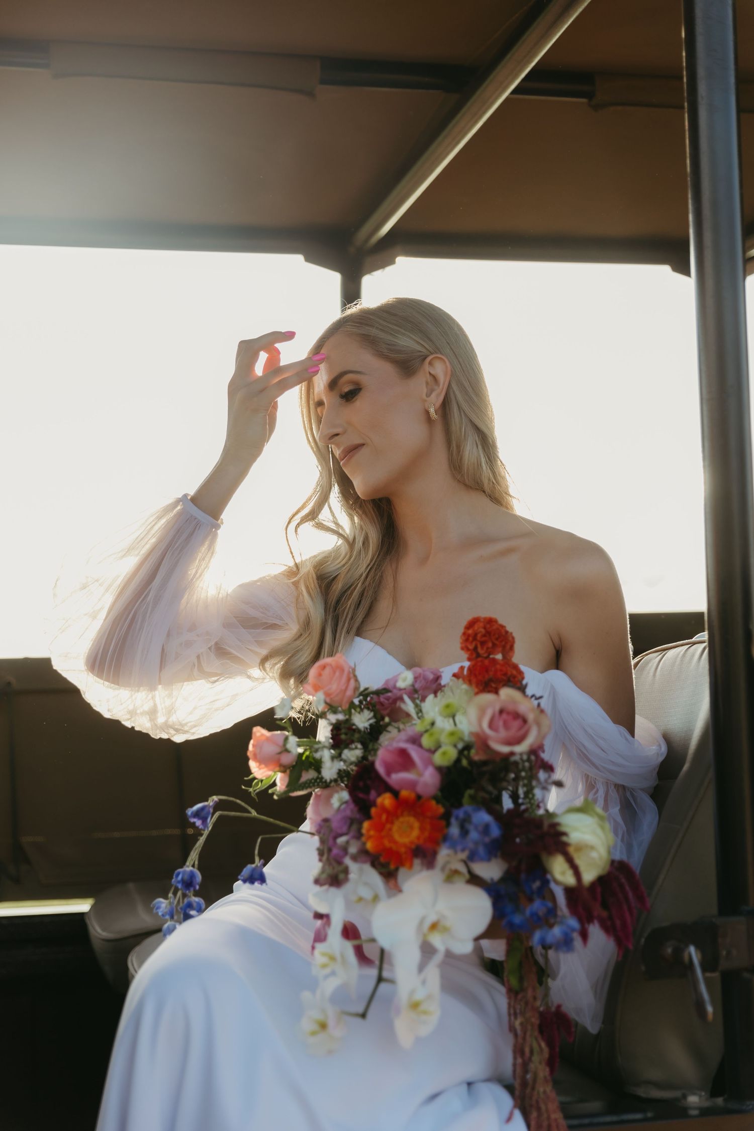Bride in white off-shoulder gown holding colorful bouquet of flowers while seated in golden sunlight.