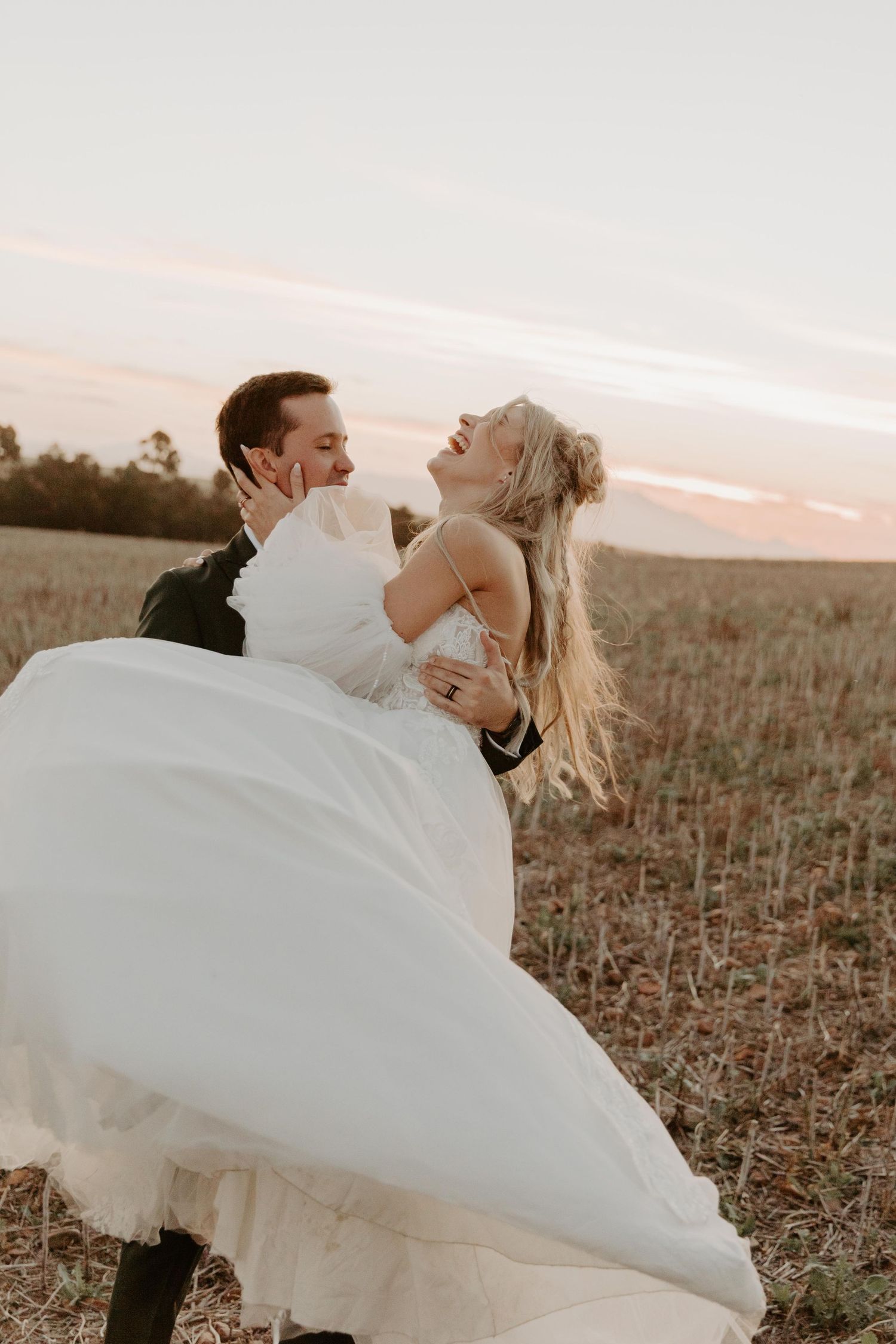 Groom lifting bride in white gown at sunset in a golden field, both laughing joyfully on their wedding day.