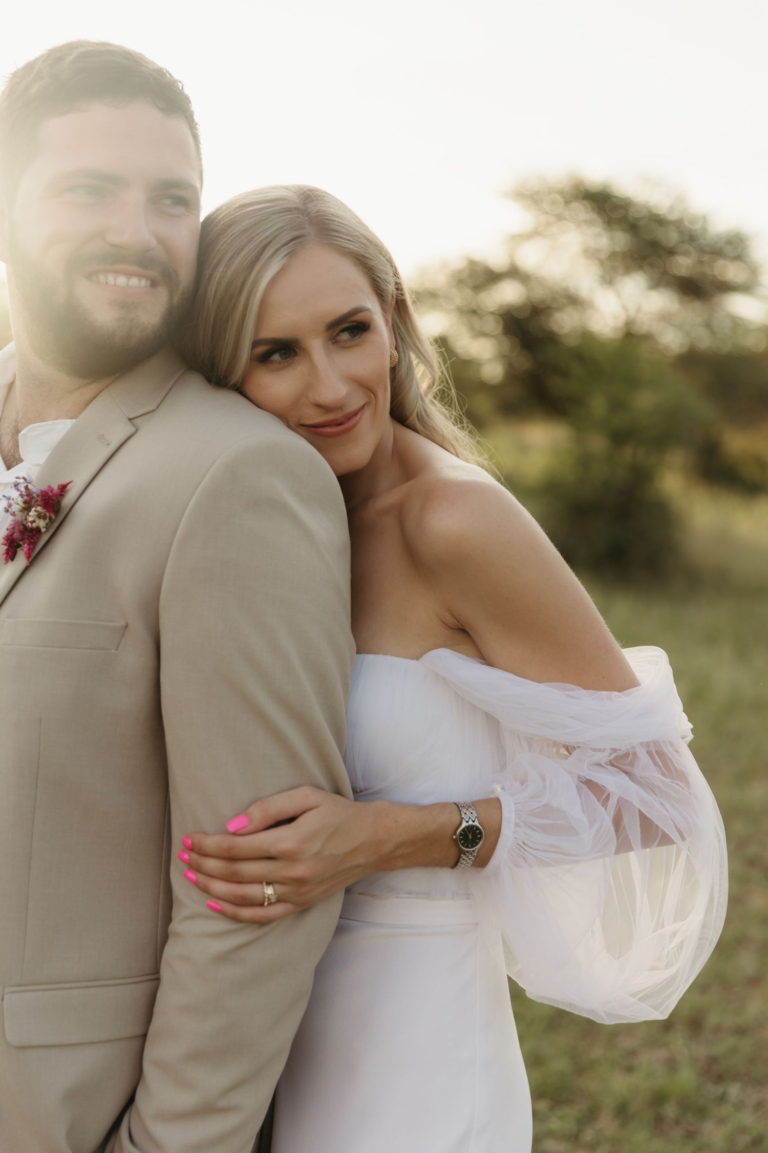 Bride in white dress embraces smiling groom in tan suit outdoors at golden hour.