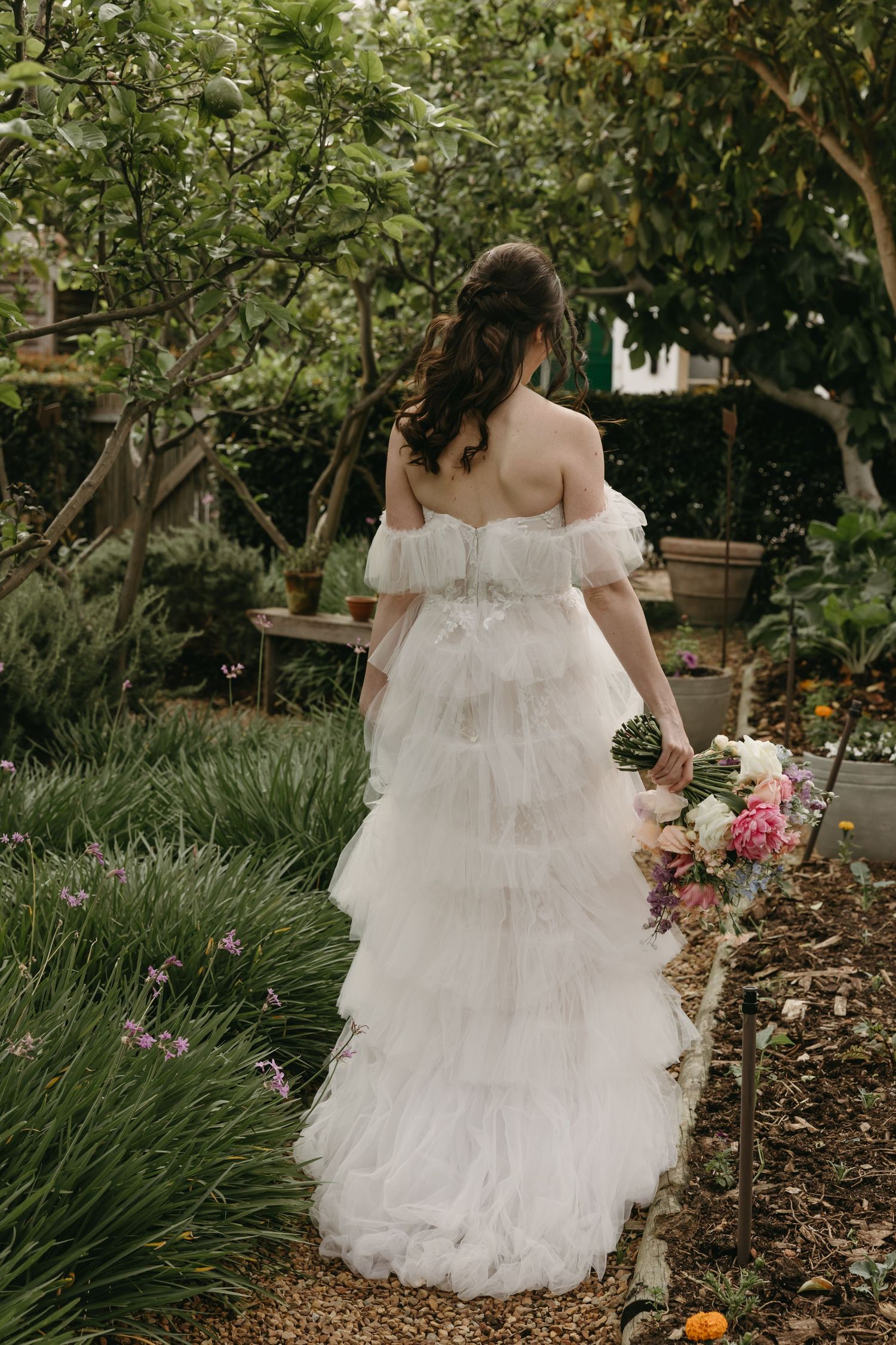 Bride in off-shoulder tiered white tulle gown holding floral bouquet, standing in lush garden path.