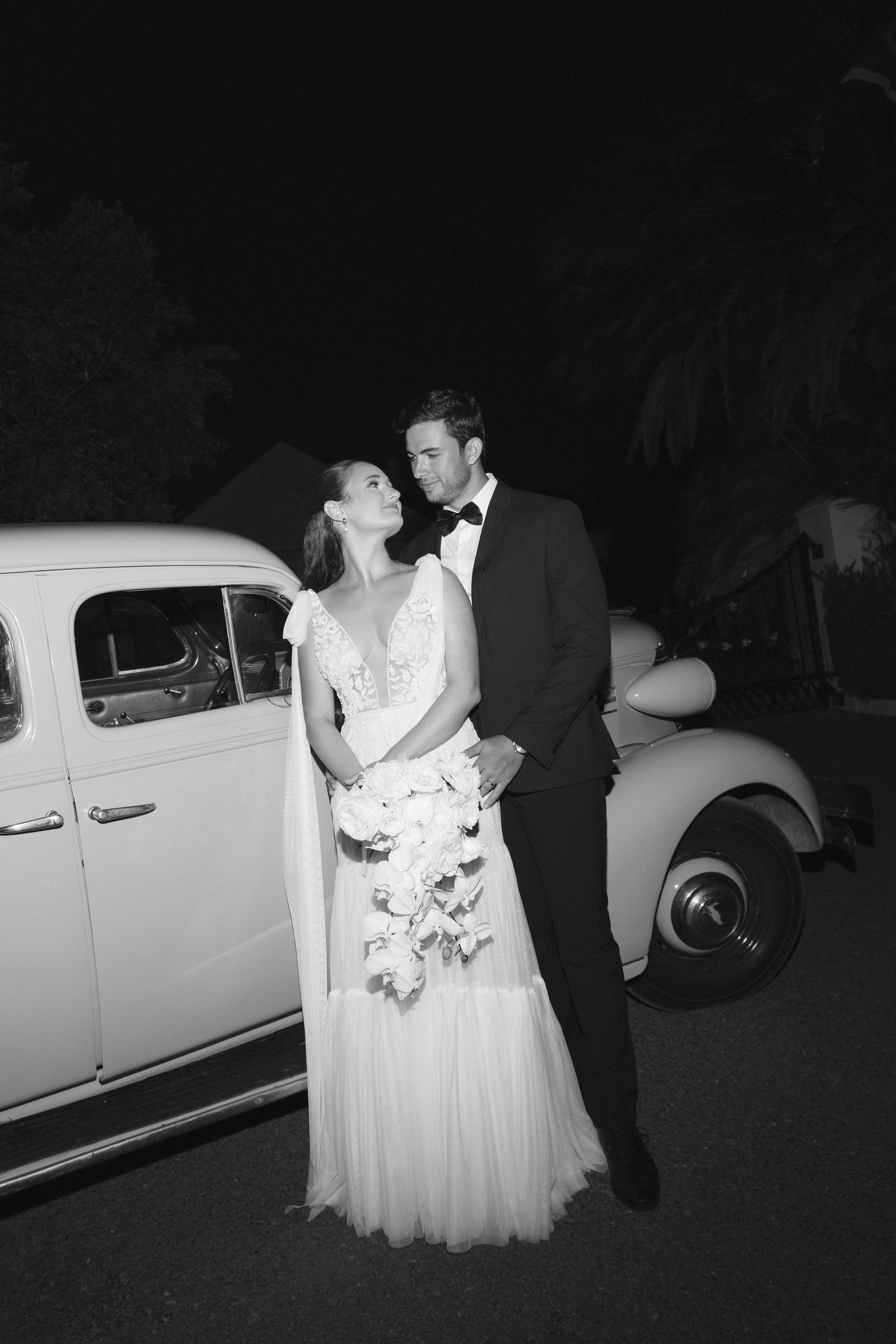 A romantic black and white wedding photo of a couple posing next to a vintage car at night.