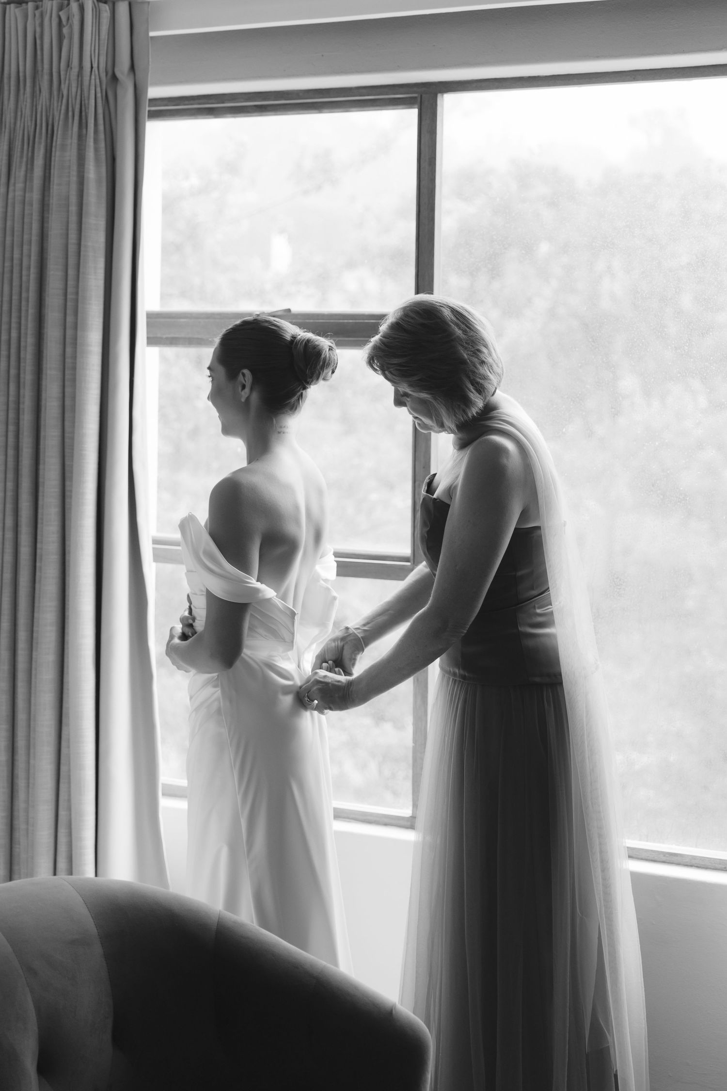Black and white photo of wedding preparation scene with bride getting dressed by window in elegant room.
