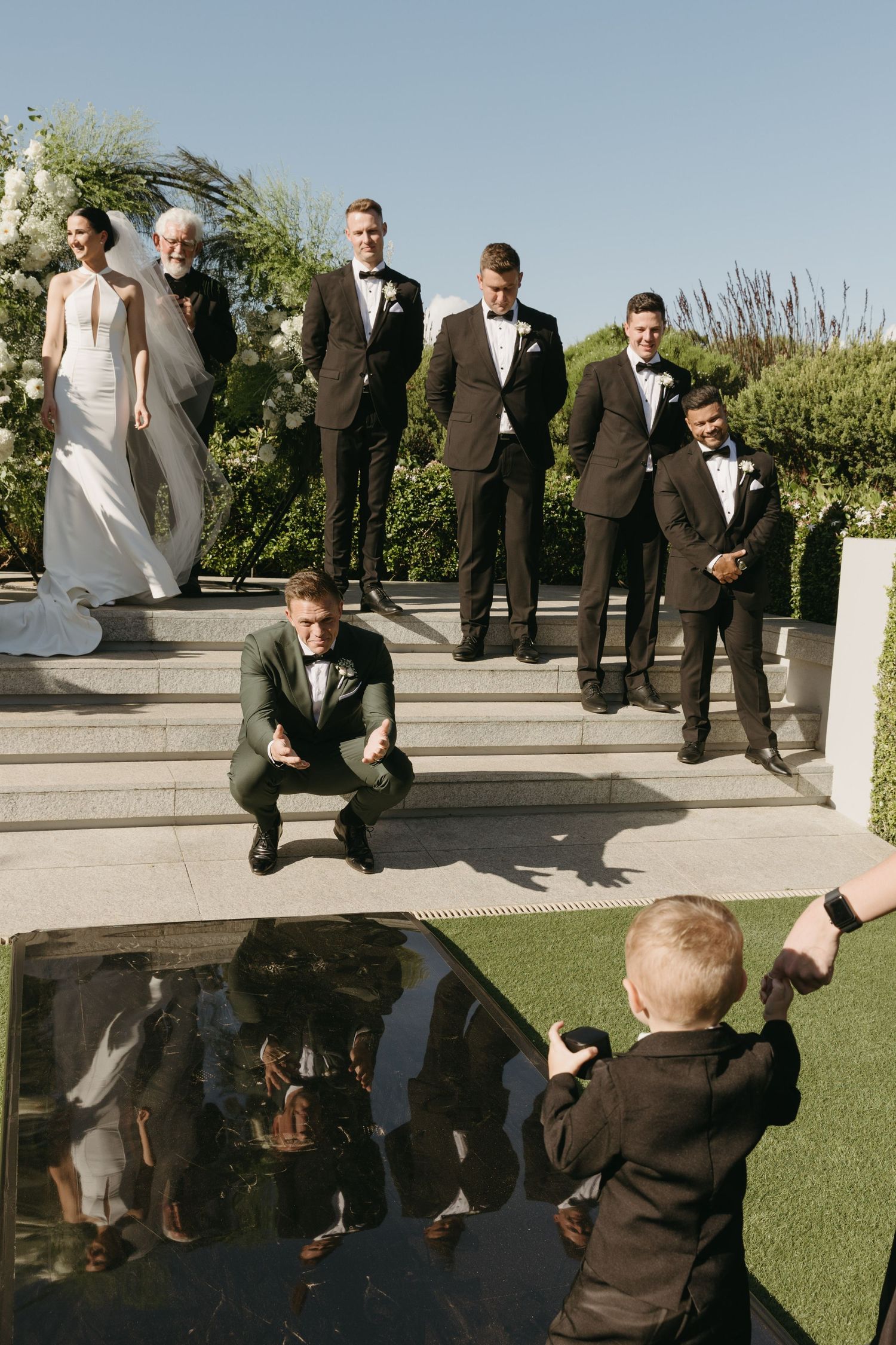 A young boy photographs a wedding party posing on outdoor steps, with the bride, groom, and groomsmen in formal attire.